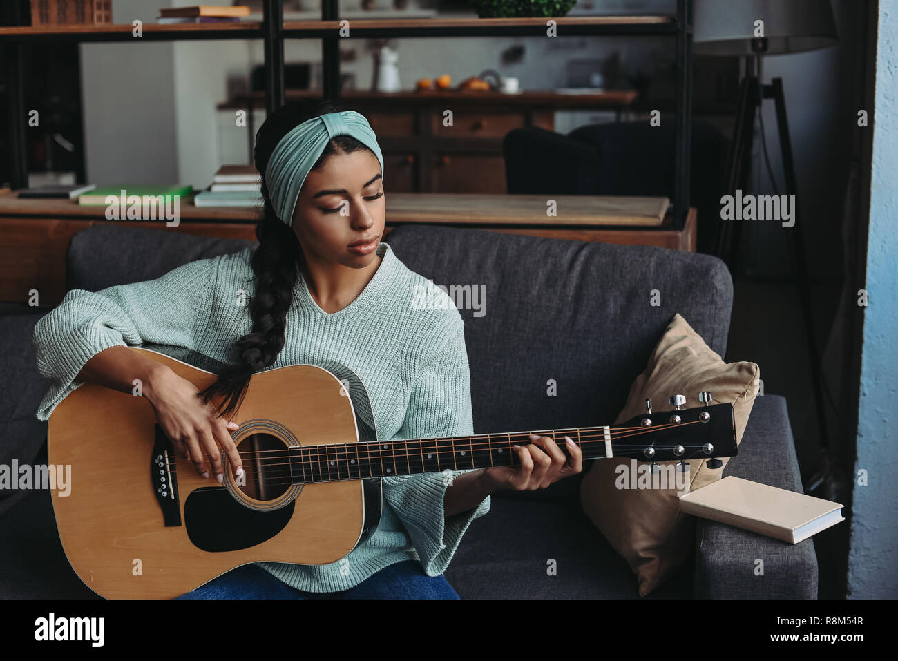 Hermosa chica de raza mixta en turquesa suéter y diadema tocando la guitarra acústica en el sofá en casa Foto de stock