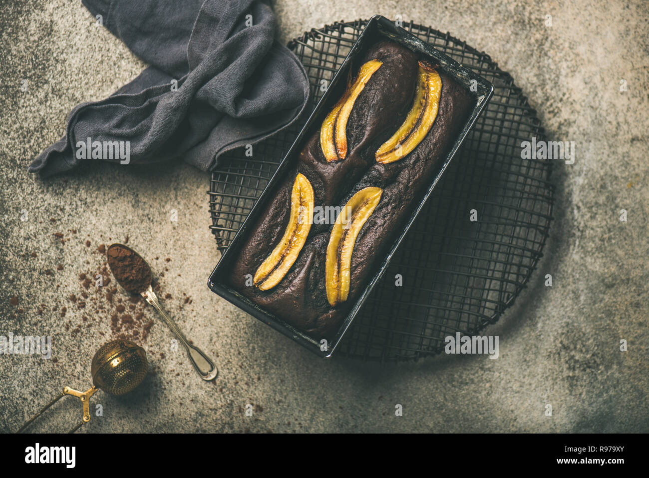 Flat-lay de chocolate recién horneadas postre torta de pan de banana en un molde para horno de rack de refrigeración con canela y cacao en polvo sobre la ficha de hormigón gris Foto de stock