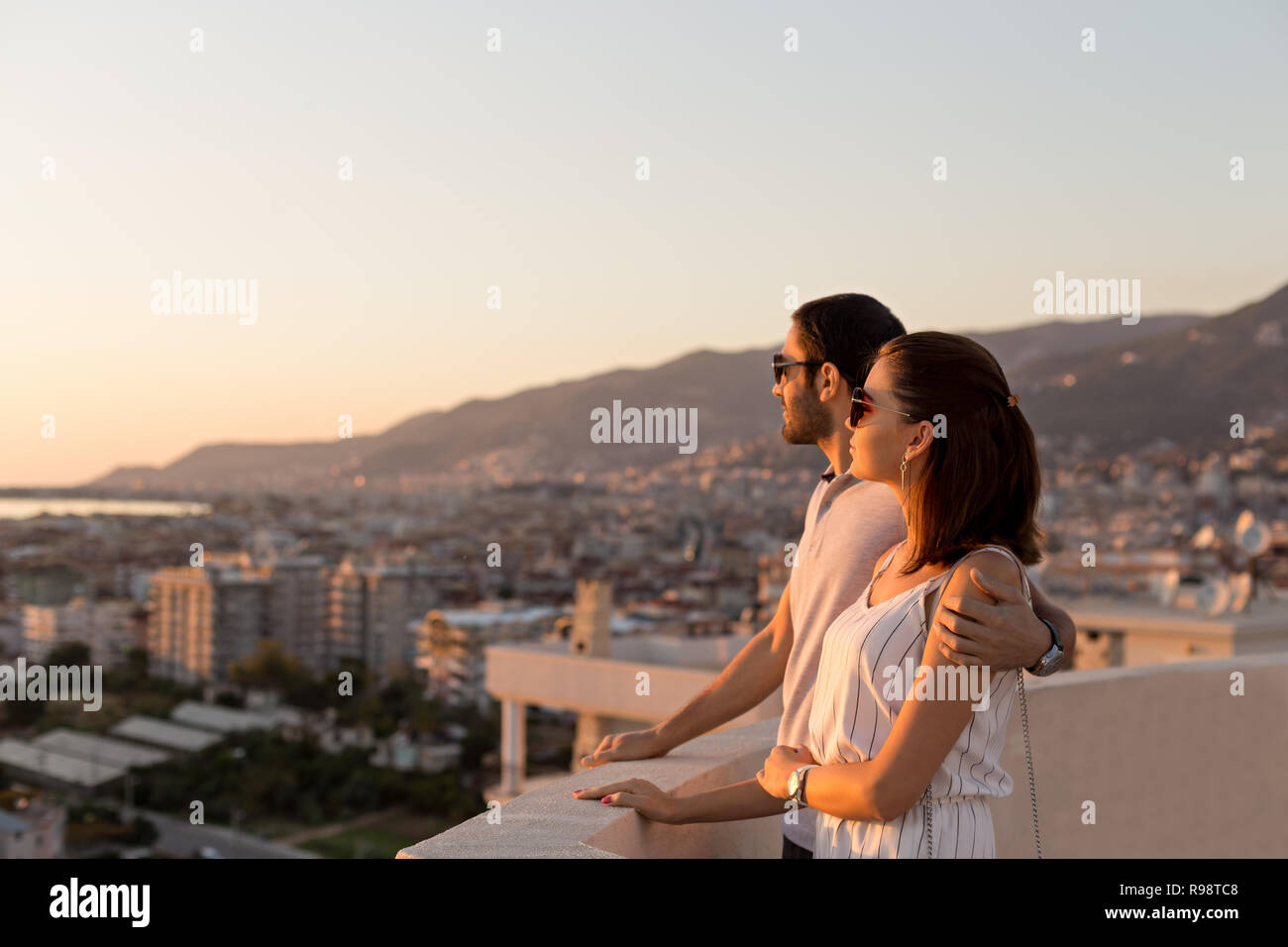 Disparo sideview horizontal de una joven pareja en atuendo de verano a disfrutar de la ciudad y vistas a la montaña desde la terraza de la azotea. Copie el espacio. Foto de stock