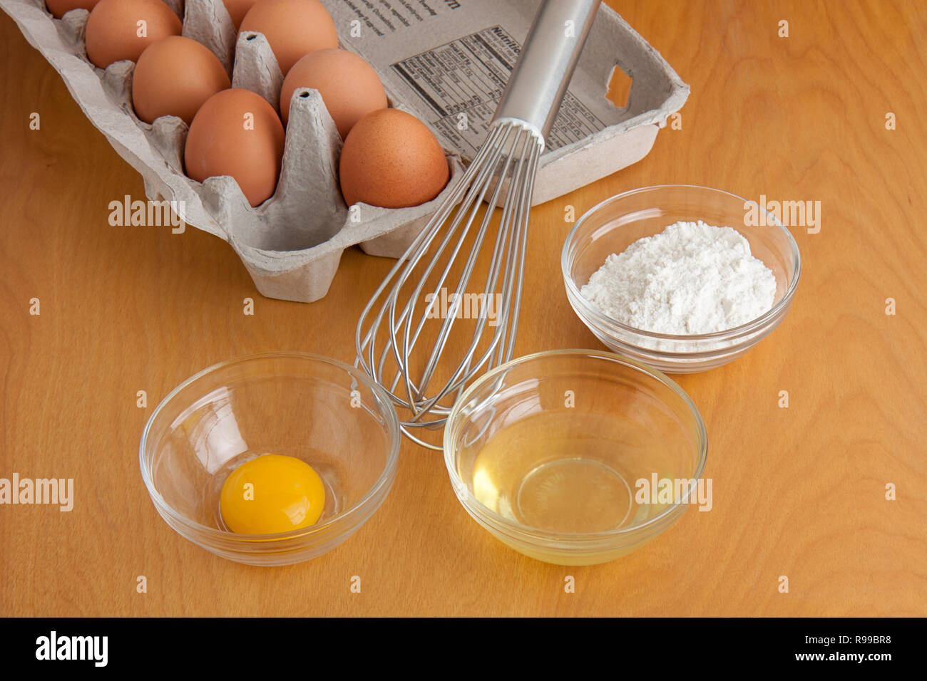 Huevos resquebrajados separados en recipientes de cristal, la harina en un tazón de vidrio, un batidor, plata y cartón de huevos todo sobre una tabla para cortar. Foto de stock