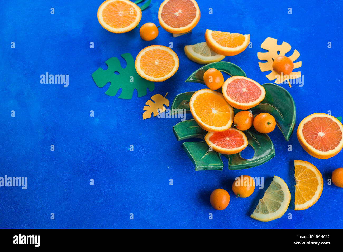 Mango, naranjas, kumquat y otras frutas tropicales en una placa de monstera. Fondo azul brillante con espacio de copia. Cabezal de comida exótica. Foto de stock