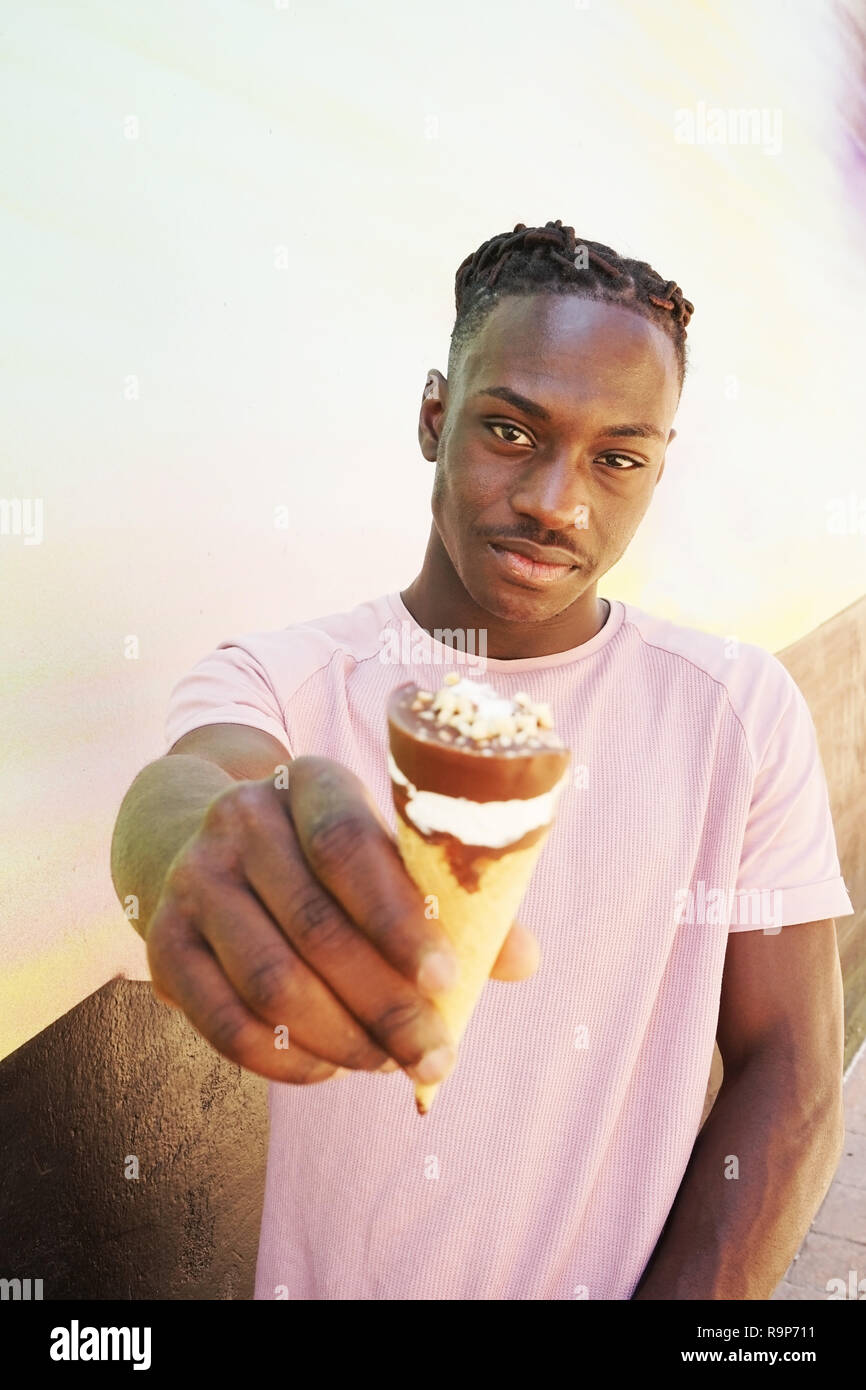 Joven guapo negro hombre viste una camiseta rosada, mantiene y se come un  cono de helado en el verano en una pintura en la pared como una puesta de  sol o día