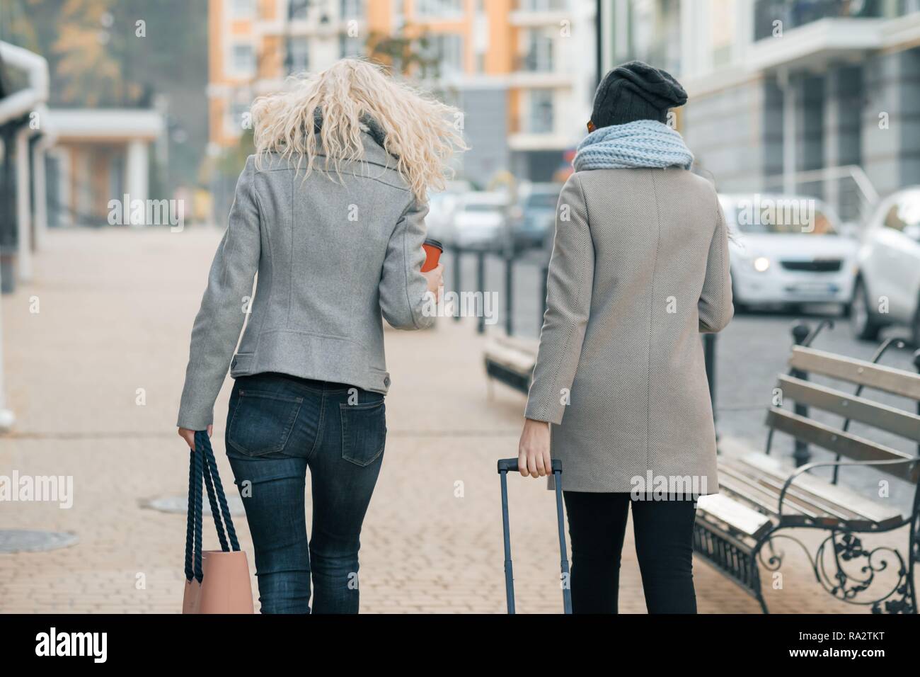Dos jóvenes mujeres hermosas en ropa de abrigo por las calles de ciudad con una de viaje, la mujer riendo y hablando, vista desde la de atrás Fotografía