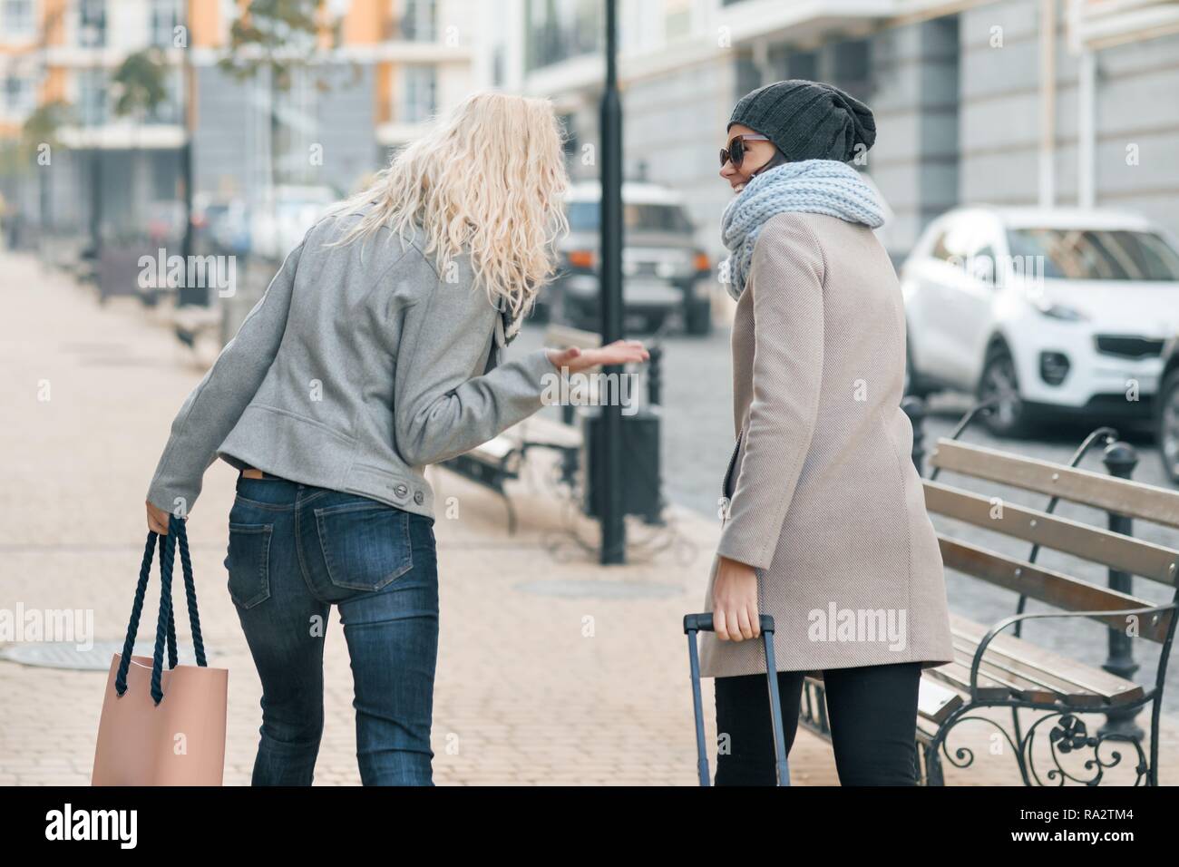 Dos jóvenes mujeres hermosas en ropa de abrigo caminando por las calles de  la ciudad con una maleta de viaje, la mujer riendo y hablando, vista desde  la parte de atrás Fotografía