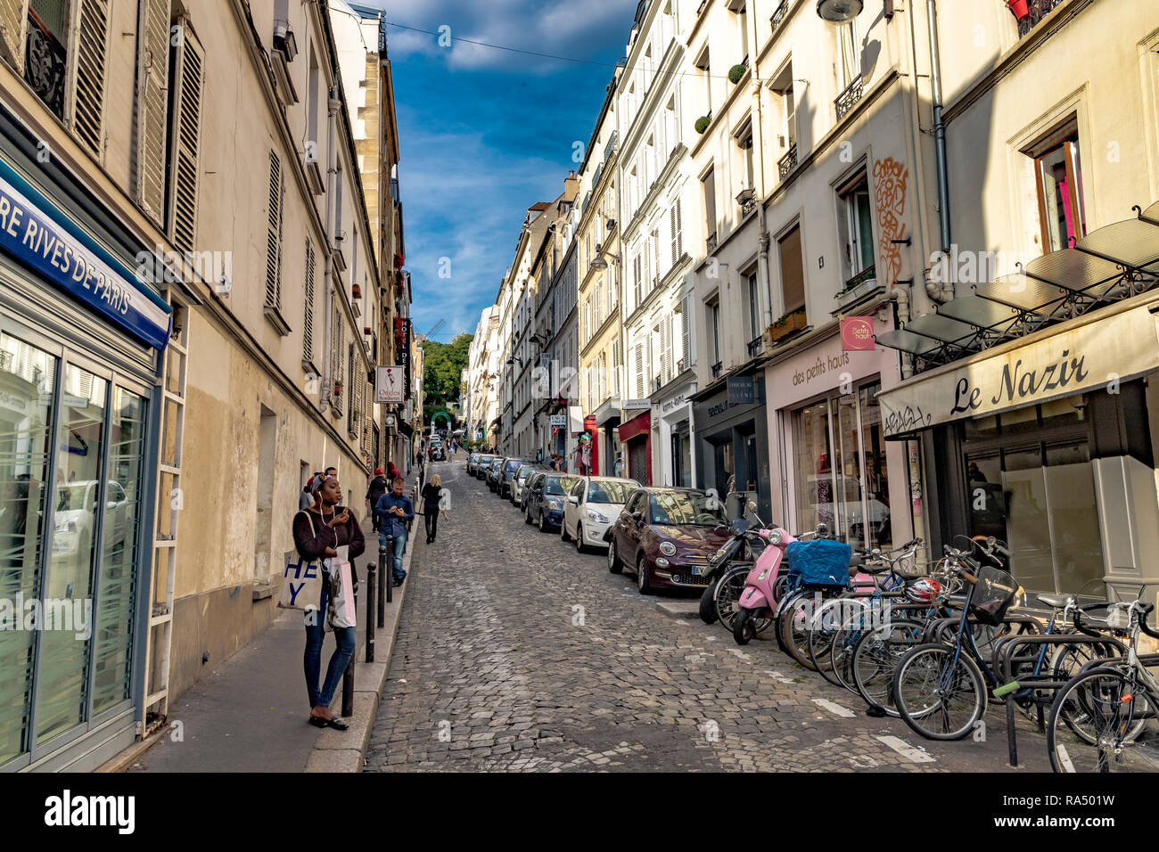 Una mujer de pie en una calle adoquinada de Montmartre con coches estacionados ,motonetas y bicicletas estacionados fuera de Café Le Nazir , Rue Tholozé ,Paris Foto de stock