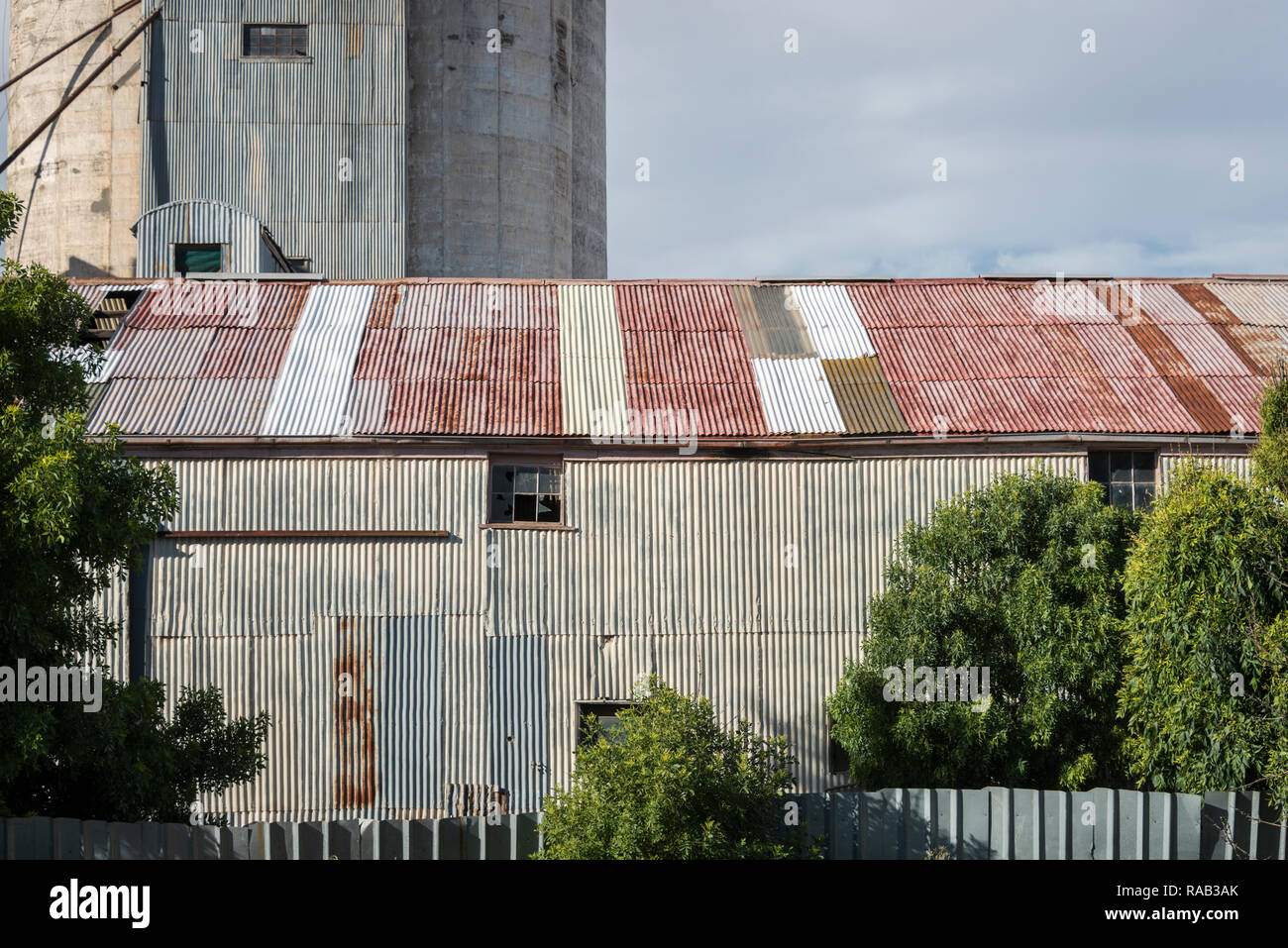 Antigua estructura ondulada con techo rojo difuminado, silo en el fondo y cielo nublado Foto de stock