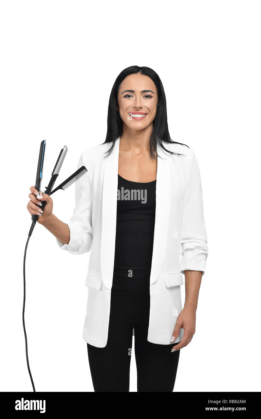 Hermosa joven peluquero buscando trabajo después de estudiar en los cursos.  Peluquería en ropa informal pero elegante celebración crest y alisadores  para el cabello en la mano. Cortadora de cabello feliz sonriente