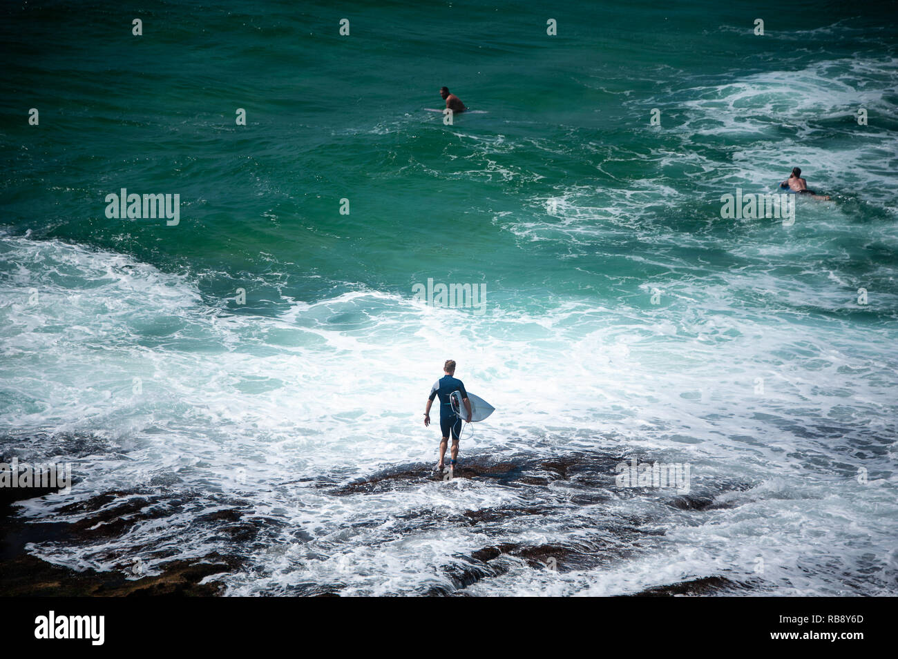 Vista aérea de un surfista llevar su tabla de surf en las olas. En traje de hombre camina en primer plano rocoso hacia un mar azul turquesa. Foto de stock