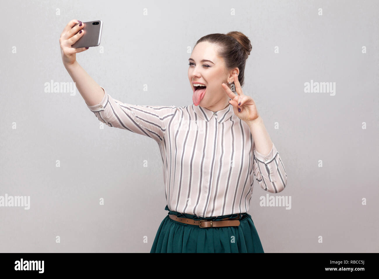 Tiempo Selfie! Retrato de feliz tonto alegre atractivo blogger mujer vistiendo en camisa a rayas de pie, guiño y mostrando la lengua y haciendo selfie Foto de stock