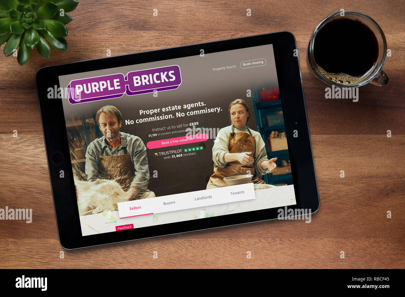 El sitio web de ladrillos color púrpura es visto en un iPad, en una mesa de madera junto con un café espresso y una planta de casa (Editorial). Foto de stock