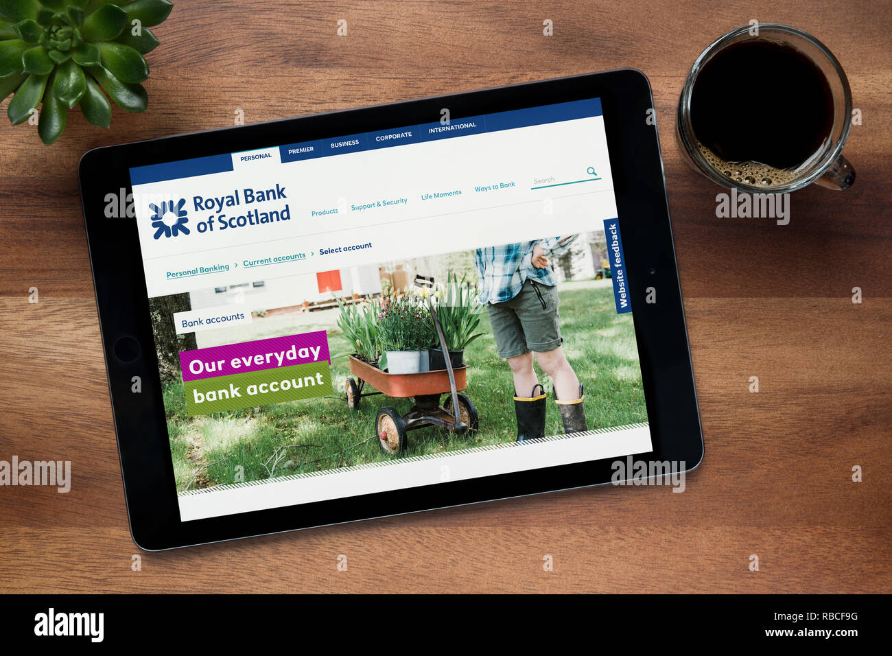 El sitio web de Royal Bank of Scotland es visto en un iPad, en una mesa de madera junto con un café espresso y una planta de casa (Editorial). Foto de stock
