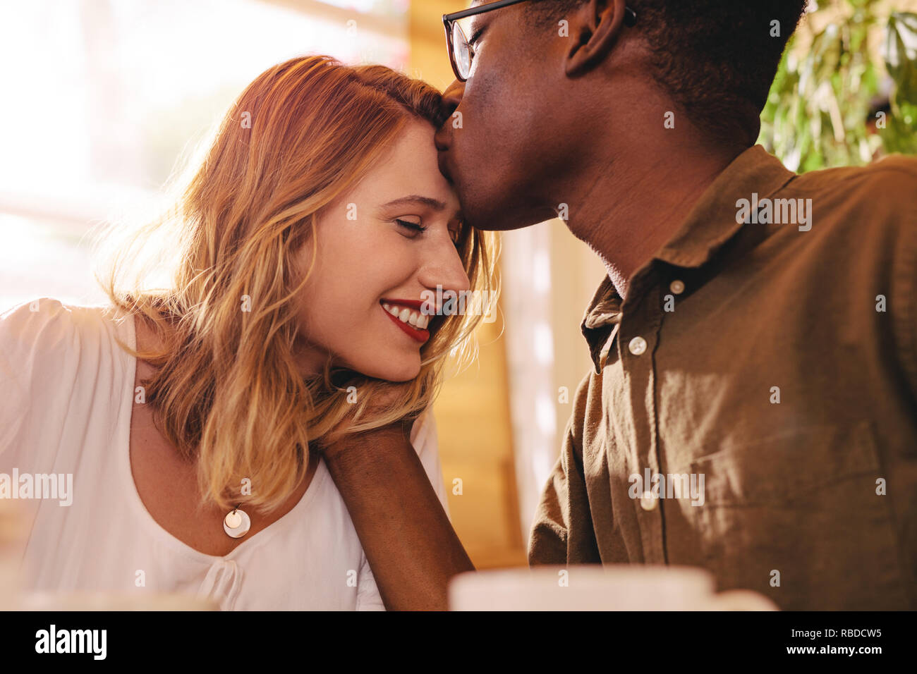 Hombre africano besar la frente de su novia. Interracial pareja amorosa en la fecha en la cafetería. Foto de stock