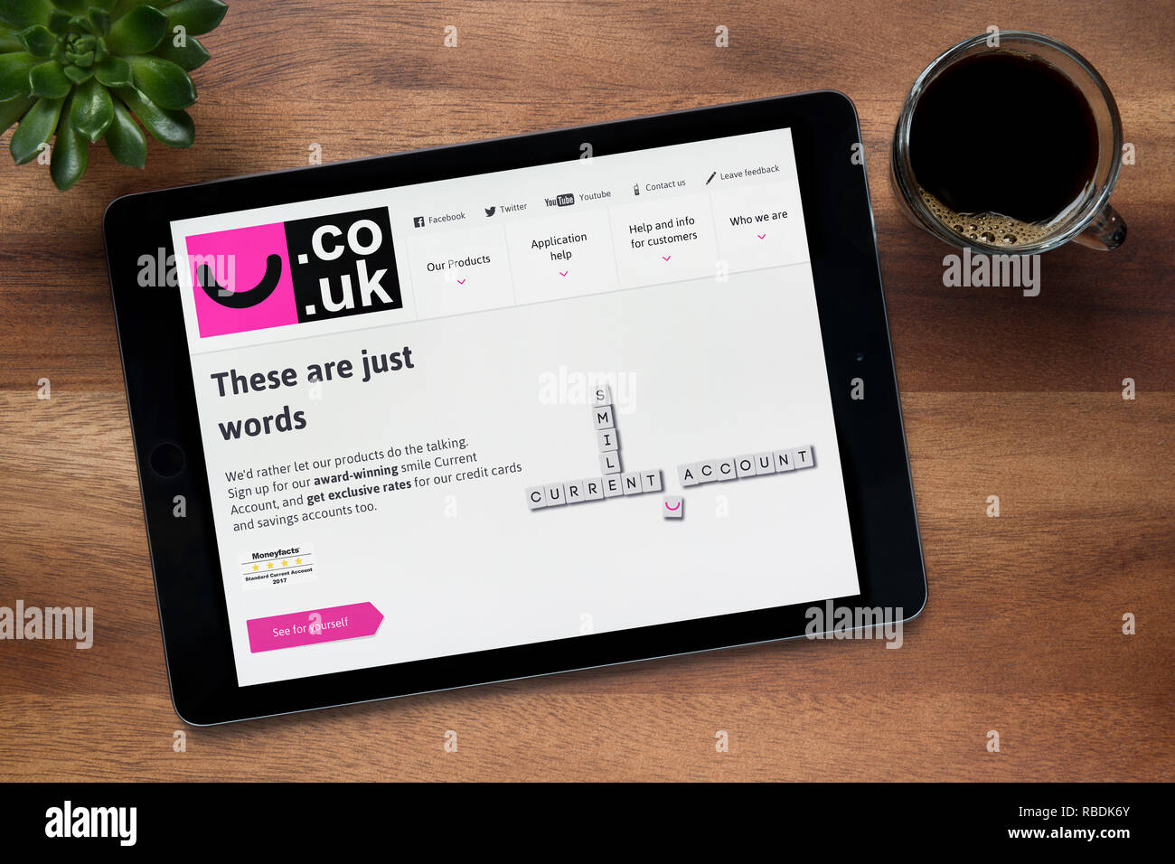 El sitio web del banco de sonrisas es visto en un iPad, en una mesa de madera junto con un café espresso y una planta de casa (Editorial). Foto de stock