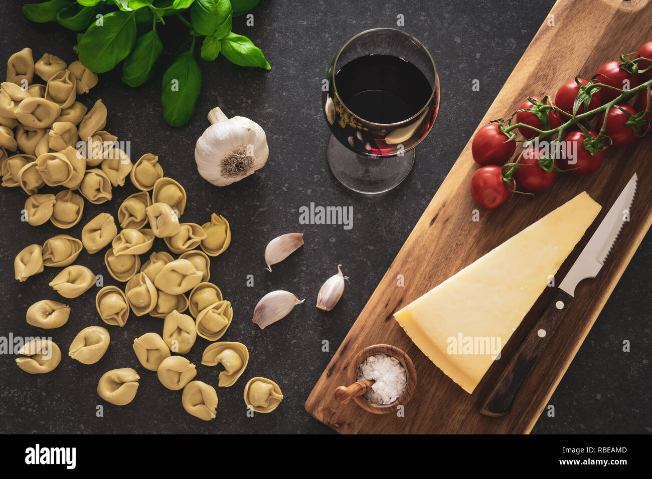 Ingredientes de comida italiana. Un vaso de vino tinto, tortellini pastas, queso y hierbas sobre fondo de piedra oscura. Foto de stock