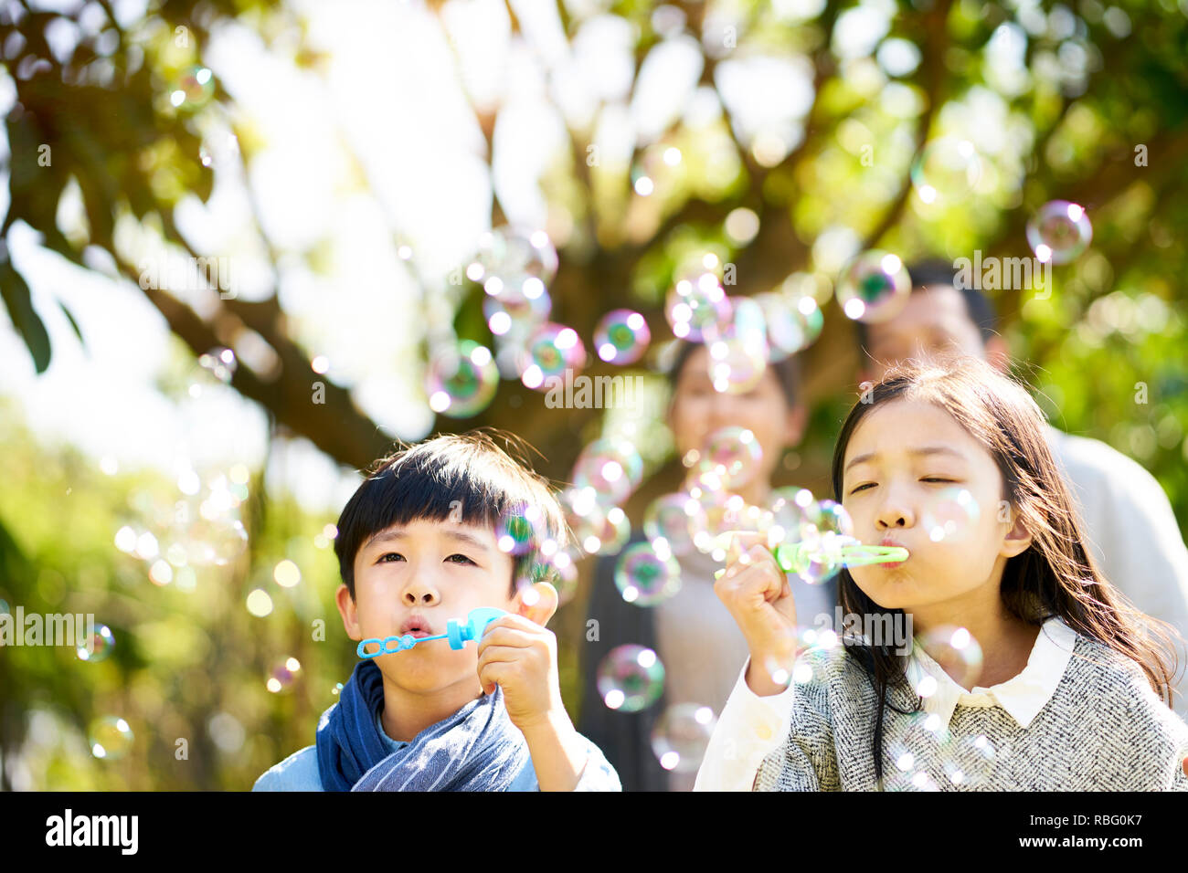 Los niños asiáticos poco chico y chica el hermano y la hermana soplando burbujas en un parque con sus padres viendo desde atrás. Foto de stock