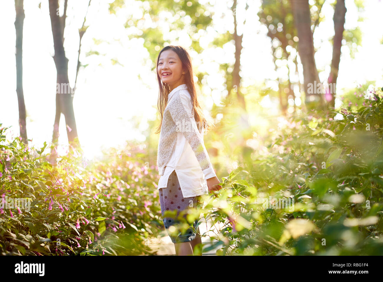 8 años de Asia pequeña y hermosa niña de pie en campo de flores mirando hacia atrás y sonriendo. Foto de stock