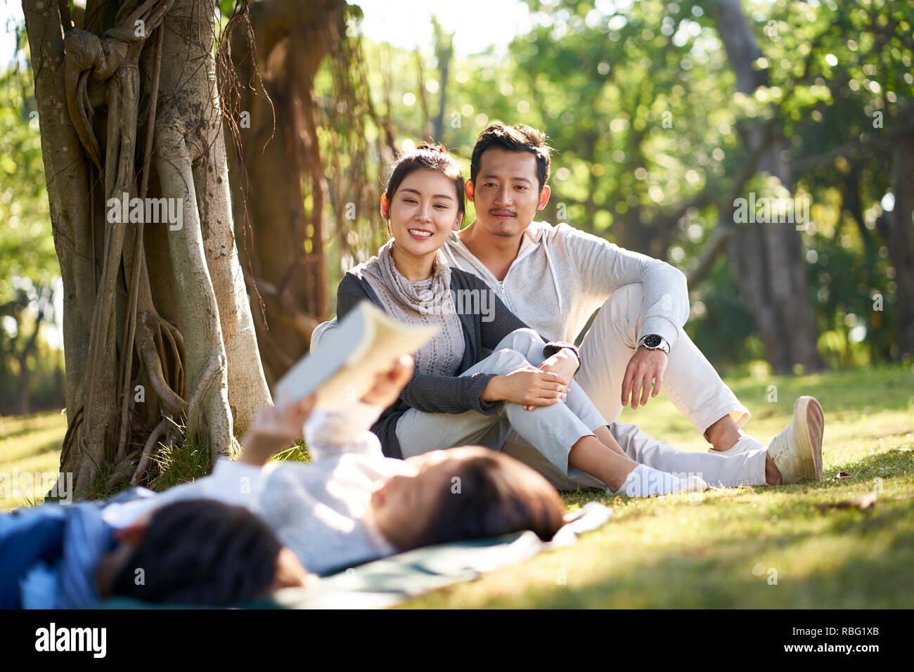 Dos niños asiáticos chico y chica divirtiéndose tumbado sobre el césped a leer un libro con sus padres sentados mirando en el fondo. Foto de stock