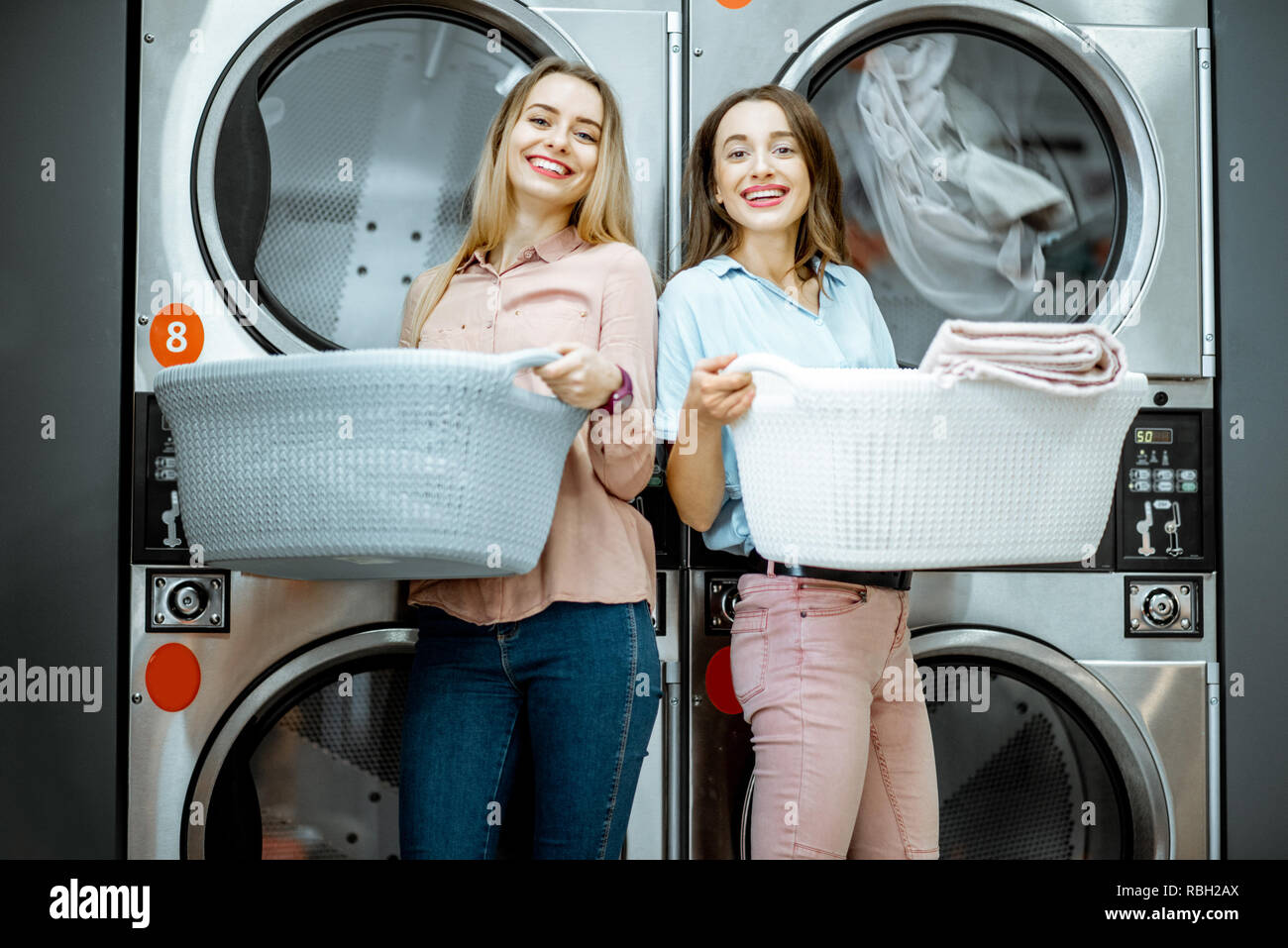 Dos jóvenes mujeres de pie junto con cestas llenas de ropa en la lavandería con secadora máquinas en fondo Fotografía de stock - Alamy