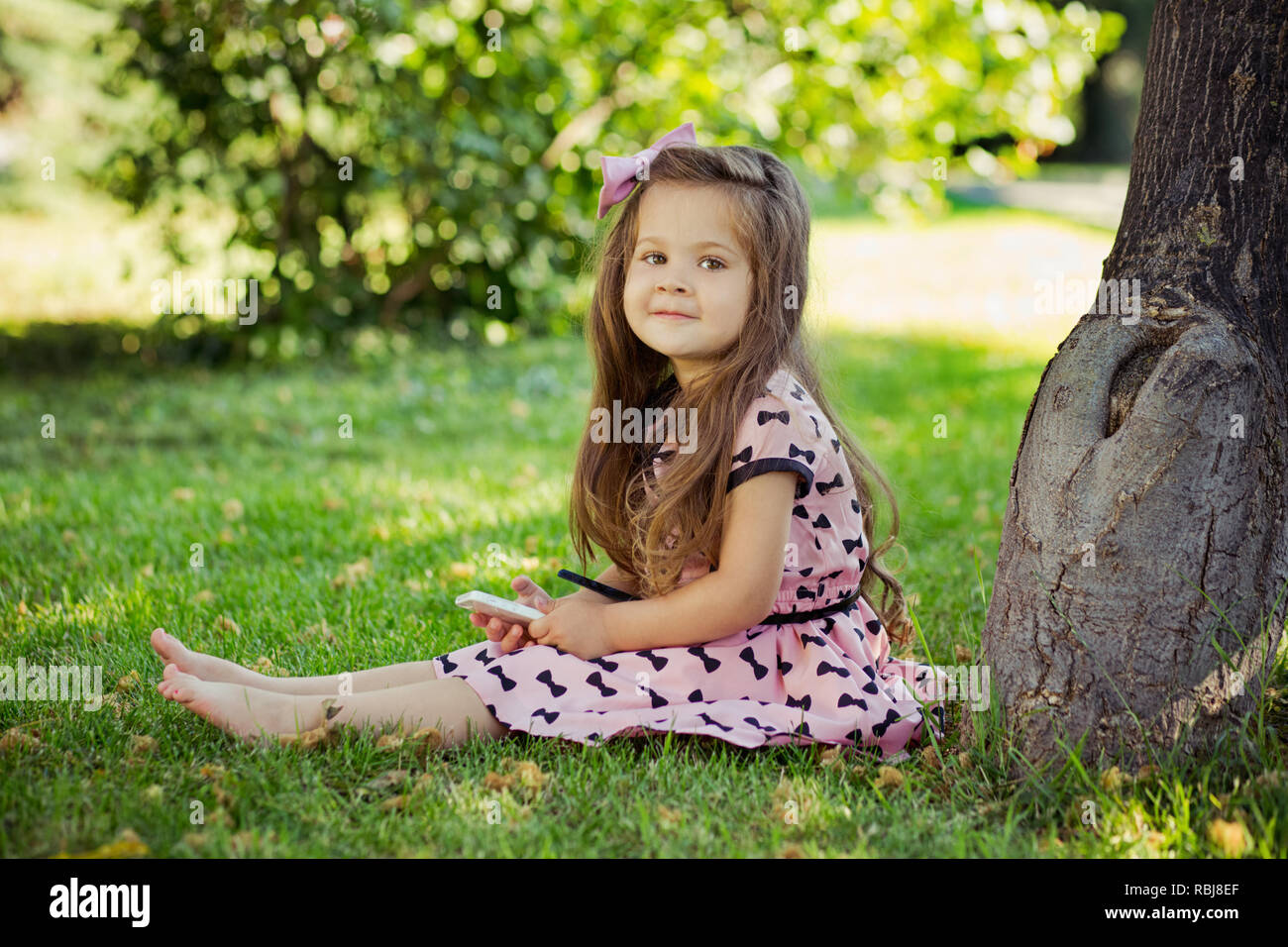Linda chica sentada en el pasto verde cerca de árbol viejo descalzo durante el verano vacaciones en awesome elegante vestido rosado con teléfono celular en su pequeña mano Fotografía de stock - Linda chica sentada en el pasto verde cerca de árbol viejo descalzo durante el verano vacaciones en awesome elegante vestido rosado con teléfono celular en su pequeña mano Fotografía de stock -
