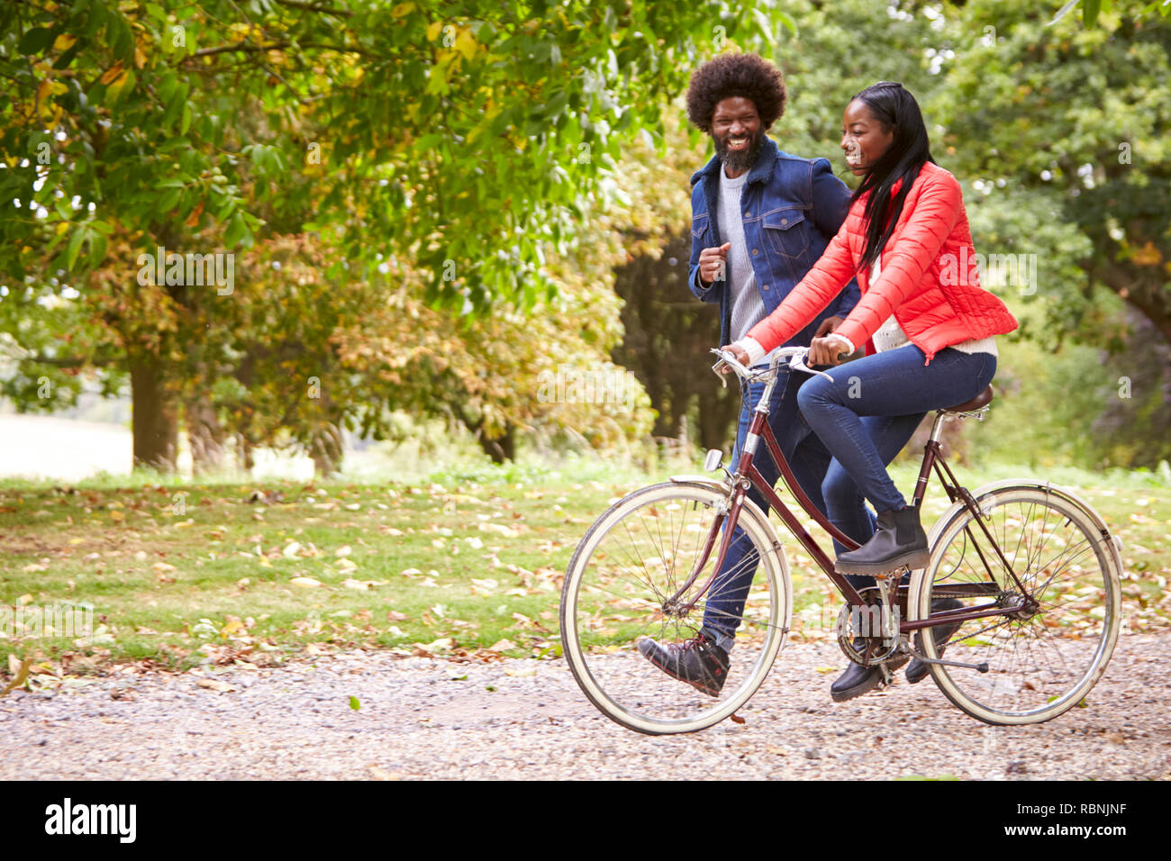 Negro corriendo en un parque junto a su novia, quien está montando una bicicleta, vista lateral Foto de stock