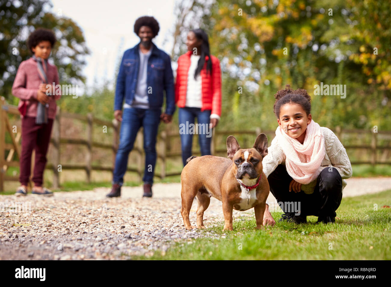 Niña de raza mixta en cuclillas a su perro mascota durante un paseo en familia en el campo, mirando a la cámara, bajo el ángulo Foto de stock