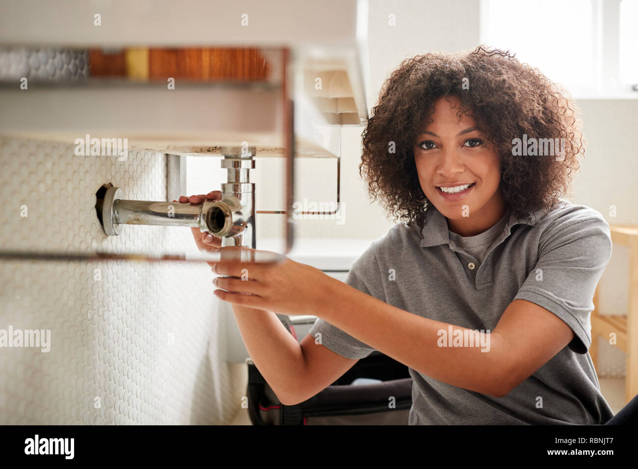 Fontanero hembra negro joven sentada en el suelo arreglar un lavabo del baño, mirando a la cámara Foto de stock