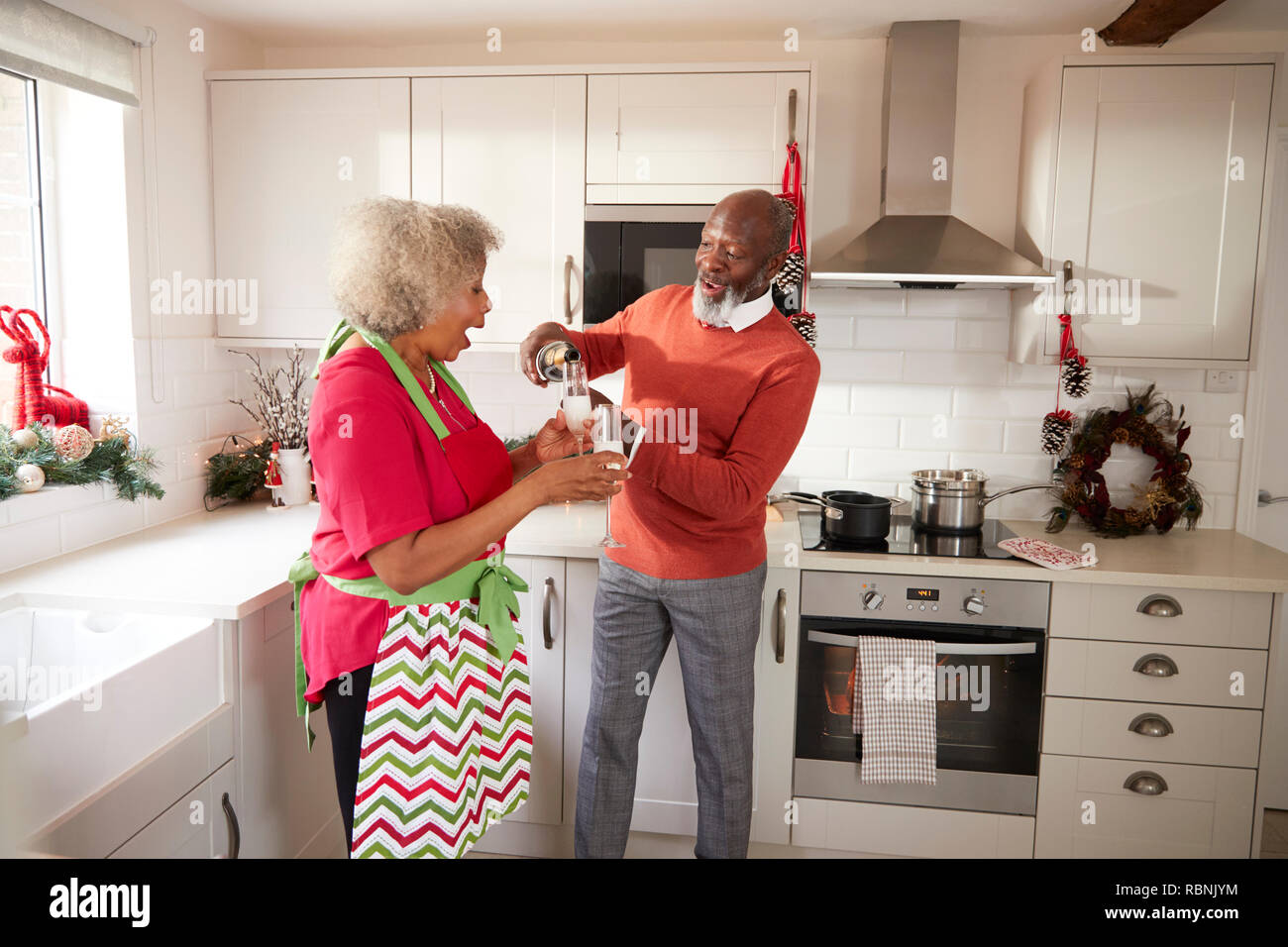 Hombre negro Senior verter champagne a su compañero en la cocina mientras prepara la cena el día de Navidad, vista lateral Foto de stock