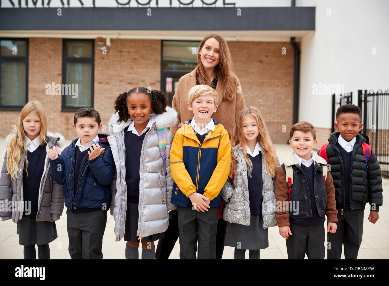 Niños de escuela primaria de pie delante de la escuela con su profesor mirando a la cámara, vista frontal Foto de stock