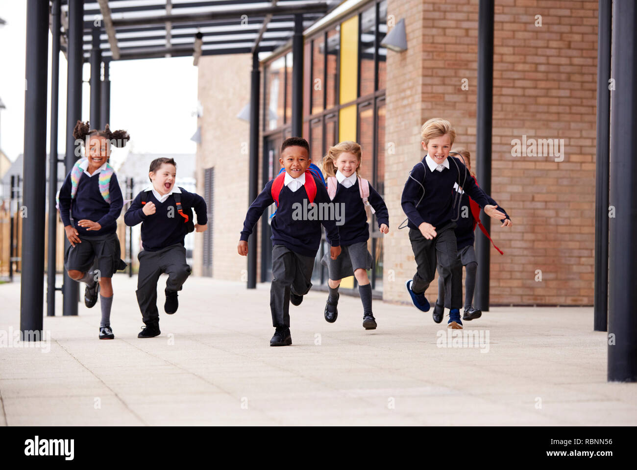 Niños de escuela primaria, vistiendo uniformes escolares y mochilas, corriendo sobre una pasarela exterior el edificio de su escuela, vista frontal Foto de stock