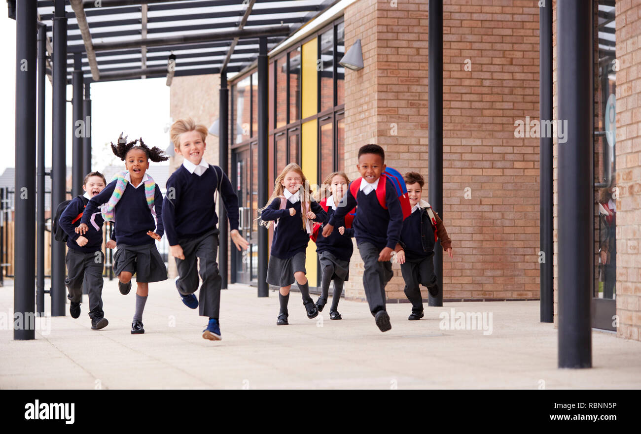 Niños de escuela primaria, vistiendo uniformes escolares y mochilas, corriendo sobre una pasarela exterior el edificio de su escuela, vista frontal Foto de stock