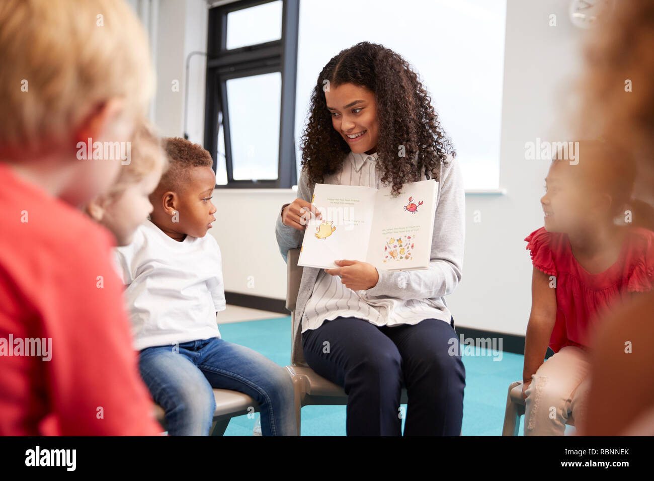 Joven maestro mostrando una imagen de un libro para niños de kinder sentadas en sillas en un círculo en el aula, de cerca, el enfoque selectivo Foto de stock