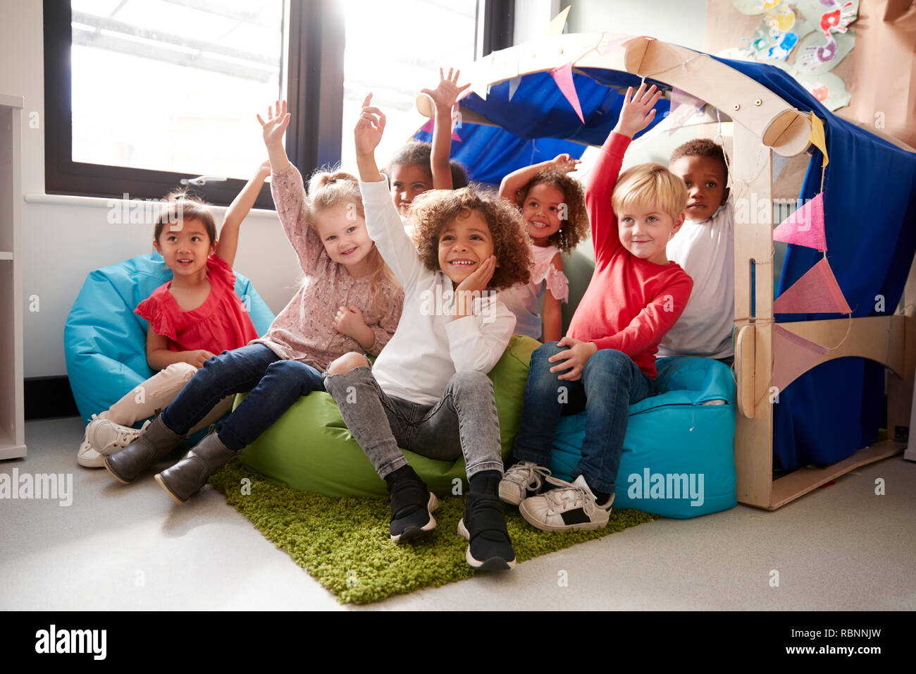 Un grupo multiétnico de escuela infantil niños sentados en sacos de frijoles en un confortable rincón del aula, levantar la mano para contestar una pregunta, bajo ángulo, cerrar Foto de stock