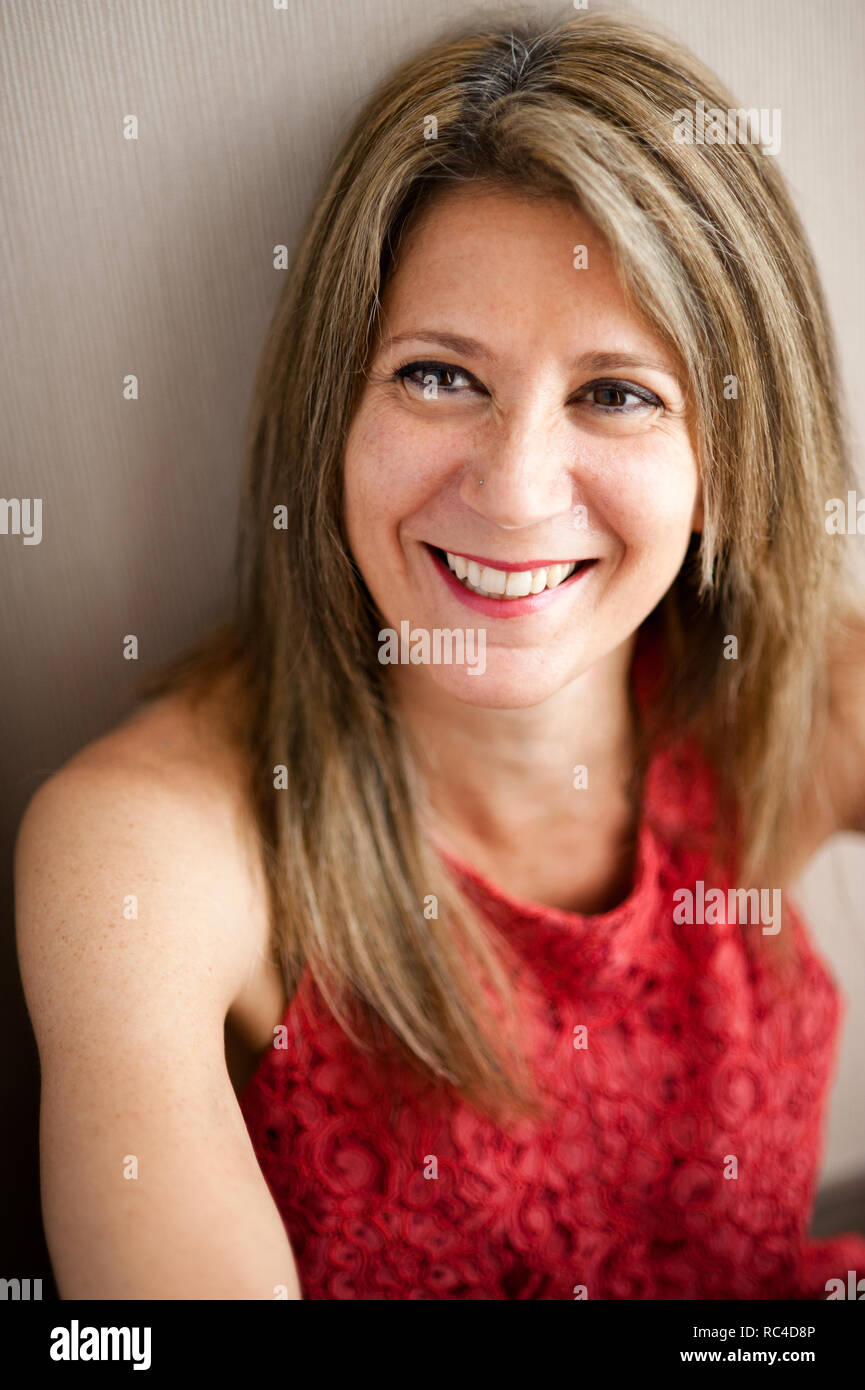 Busto retrato de mujer sonriente a la edad de 50 años, con el pelo rubio y maquillaje, vestidos de rojo vestido sin mangas, inclinándose hacia atrás en la pared gris, mirando lejos de ingenio Foto de stock