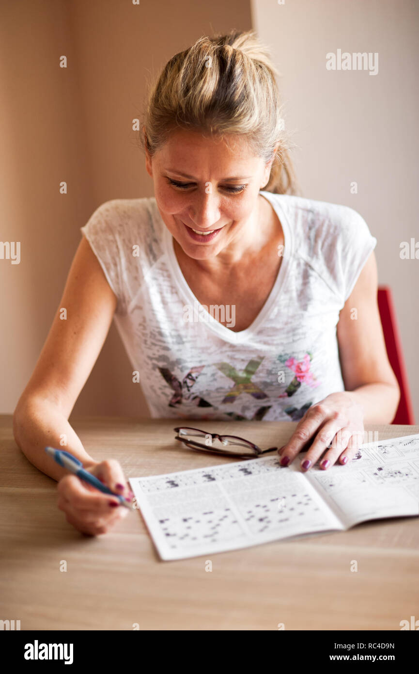 Mujer en camisa blanca sentado en la mesa de la habitación y resolver crucigramas, mirando sonriente y papel abierta Foto de stock