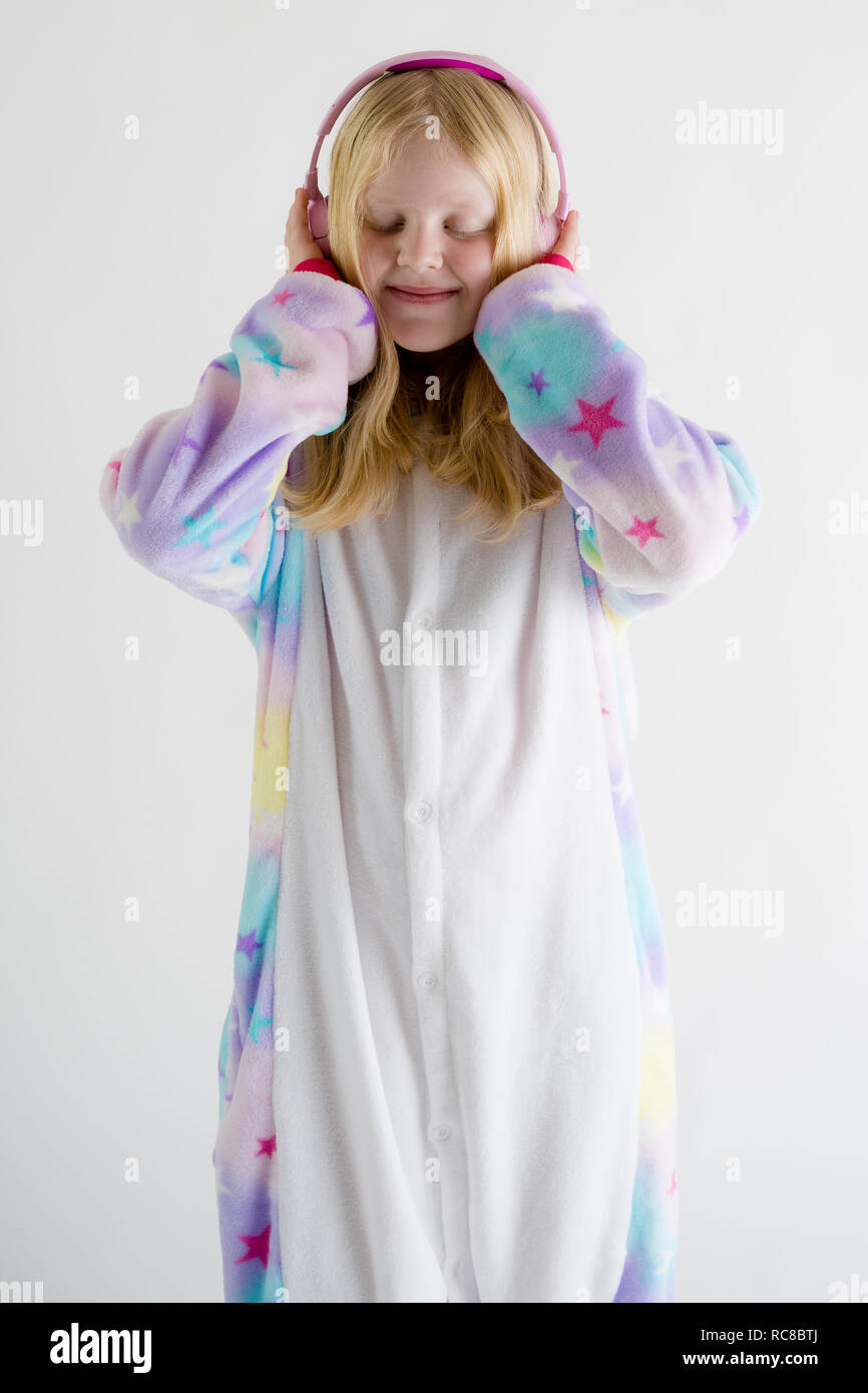 Moda moderna - hermosa chica rubia escucha música con auriculares sobre un fondo blanco en pijama kigurumi Foto de stock
