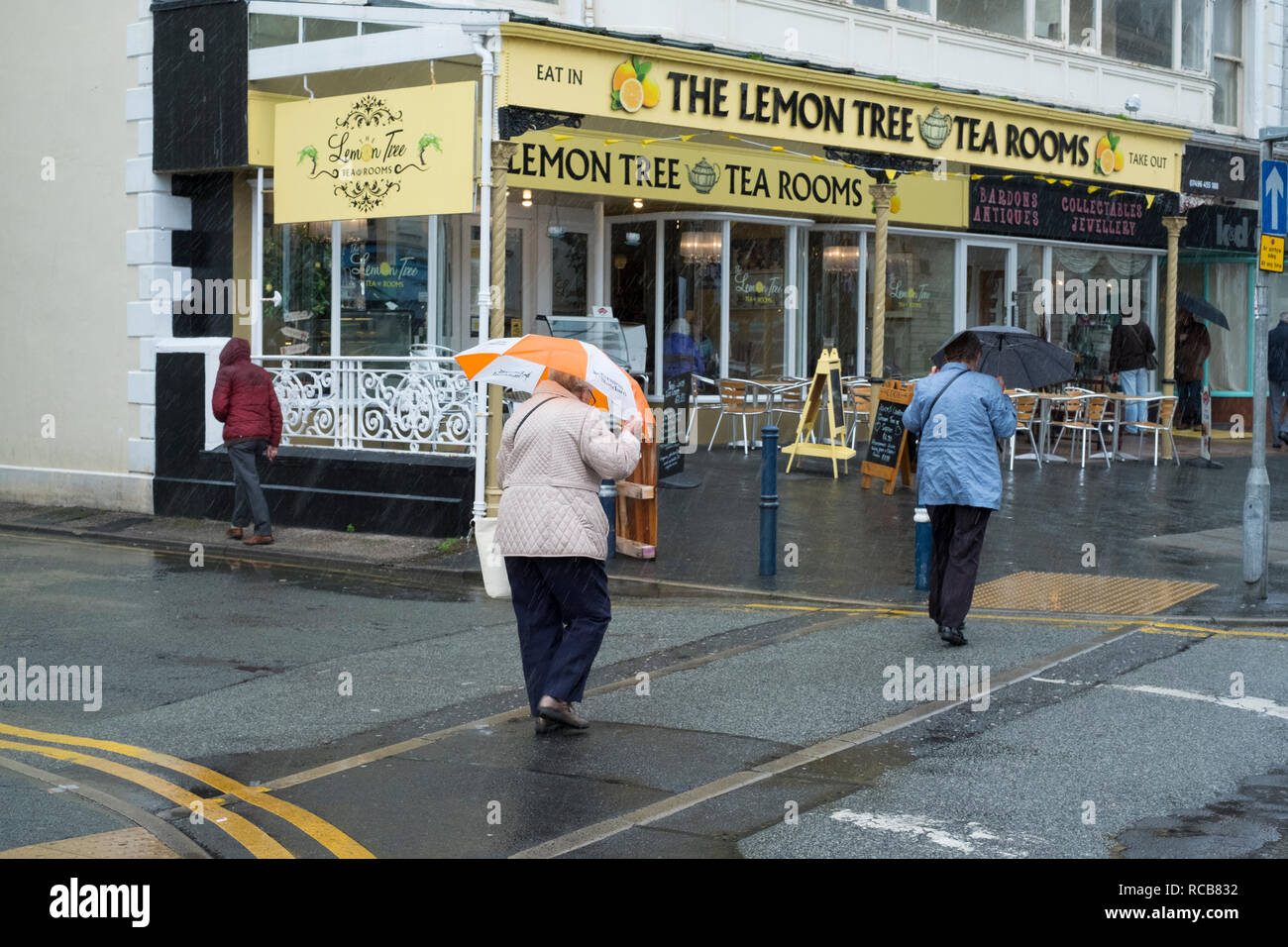 Dos mujeres con paraguas en la lluvia pasando el Lemon Tree Salones de té en Llandudno, Conwy, Gales, Reino Unido Foto de stock