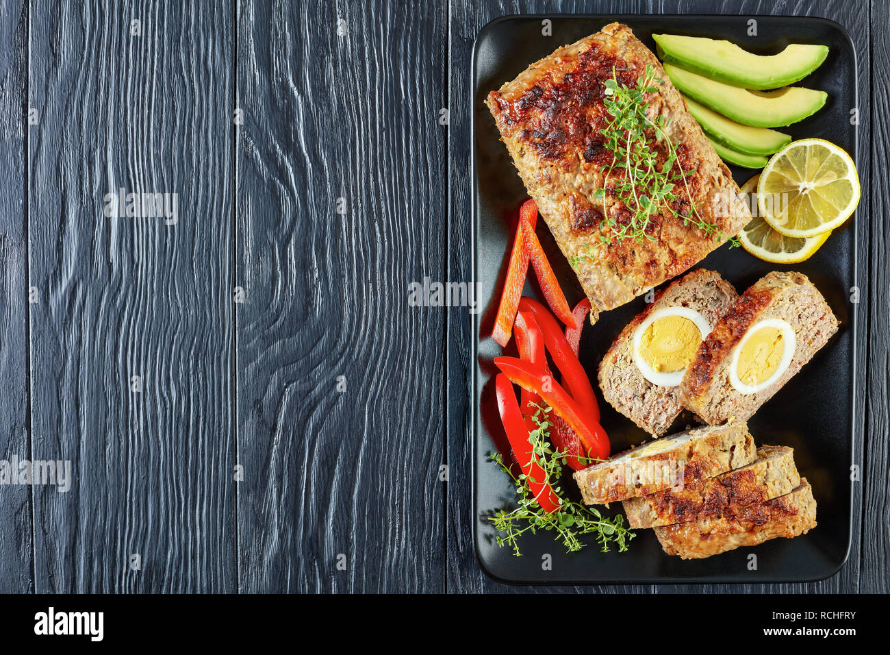 Pastel de Carne con huevo duro llenado servidos con pimiento rojo y rodajas de aguacate en un plato rectangular negra sobre una mesa de madera, vista desde arriba, fla Foto de stock