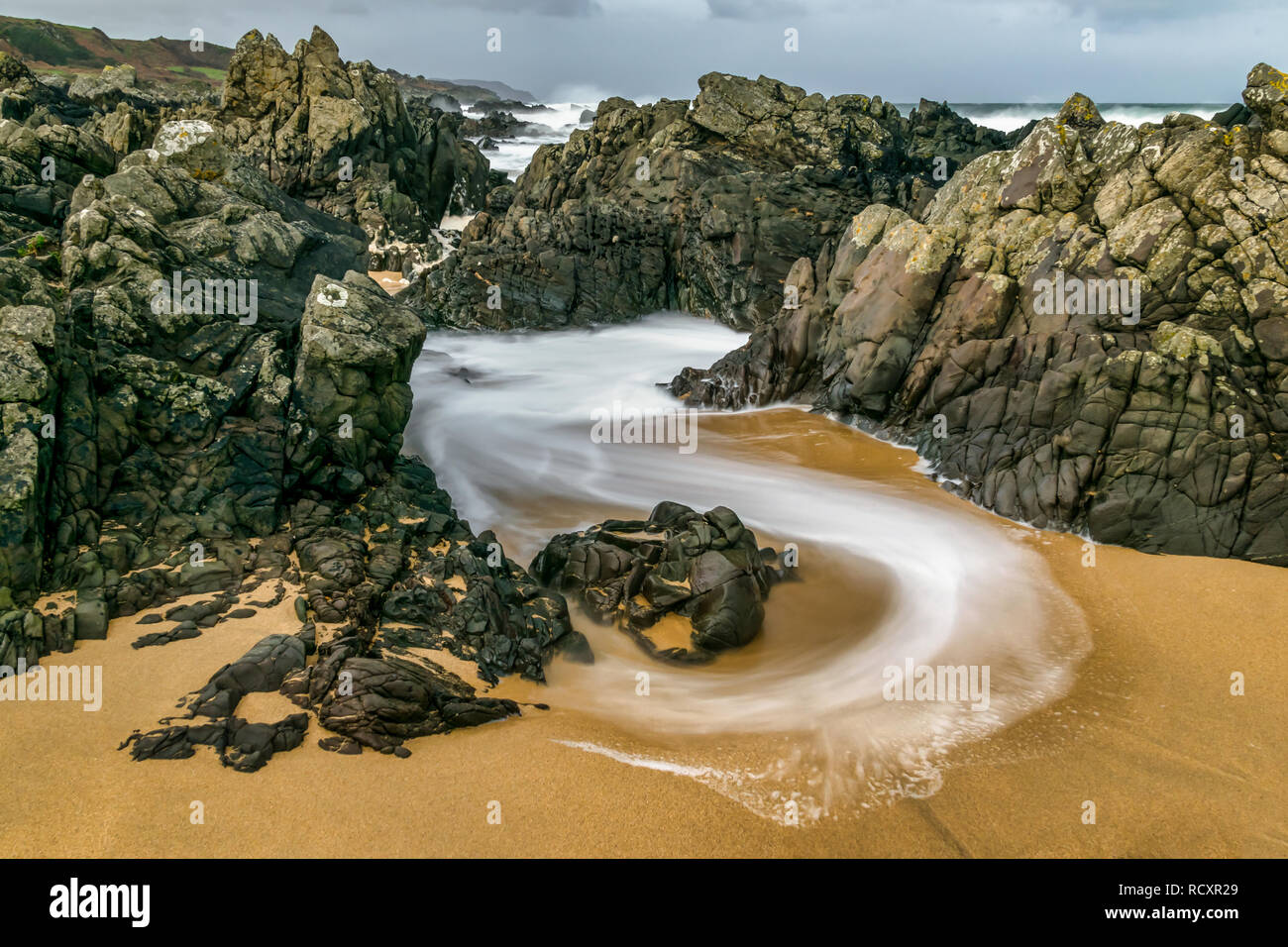 Velocidad de obturación lenta fue utilizada para captar esta imagen de un remolino de onda aunque las rocas en la playa. Al retirarse el agua crea curvas de espuma blanca Foto de stock