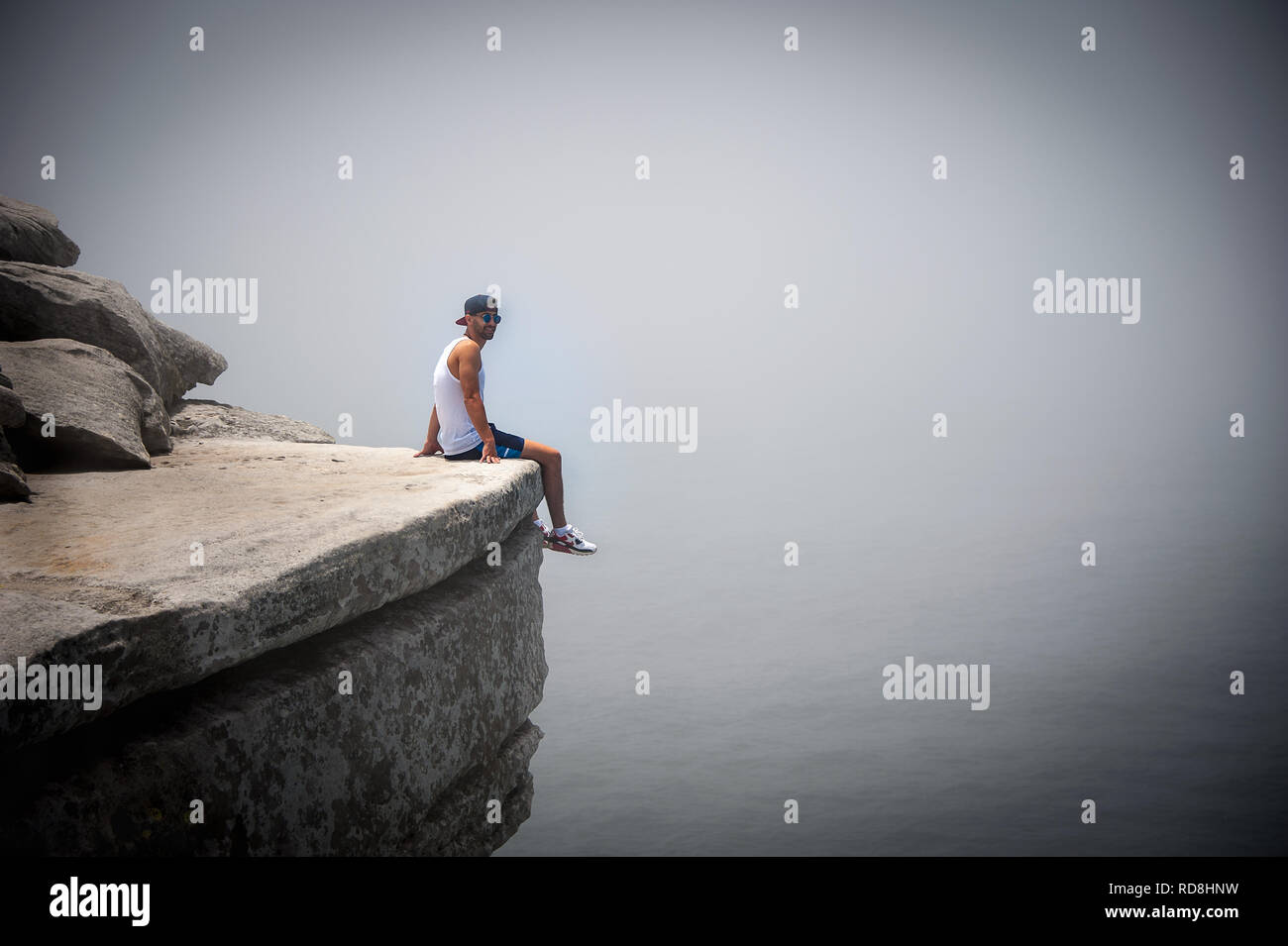 Sydney, Australia: sonriente joven sentada sobre el borde de un acantilado con caídas al océano abajo. Escena surrealista como la niebla envuelve el paisaje de mar Foto de stock