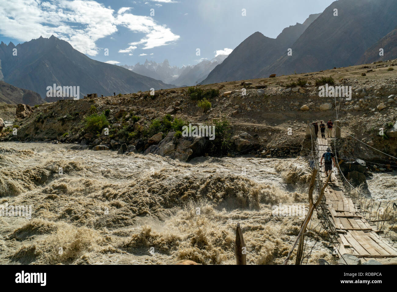 Karakoram, Pakistán - Julio 24, 2018: Aventureros cruzar el río en Puente frágil a lo largo de un río salvaje de las montañas Karakoram en Pakistán. E ilustrativos Foto de stock
