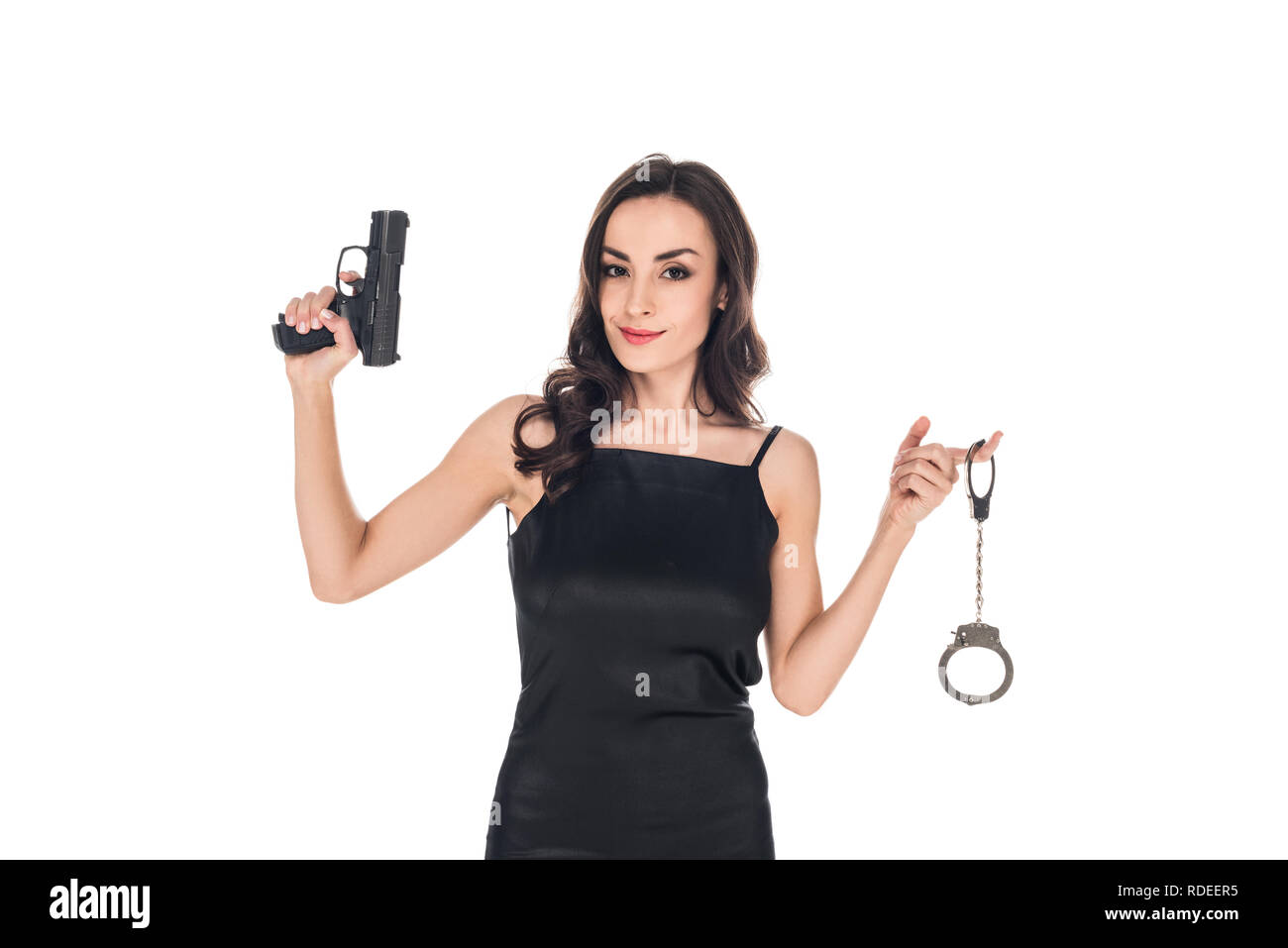 Atractivo sonriente agente secreto en vestido negro sujetando la pistola y esposas, aislado en blanco Foto de stock