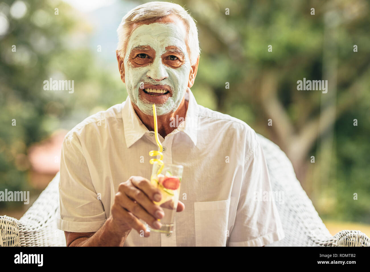Gracioso anciano con máscara de arcilla en el rostro de beber zumo. Hombre jubilado en casa cuidando de su piel en la vejez. Foto de stock