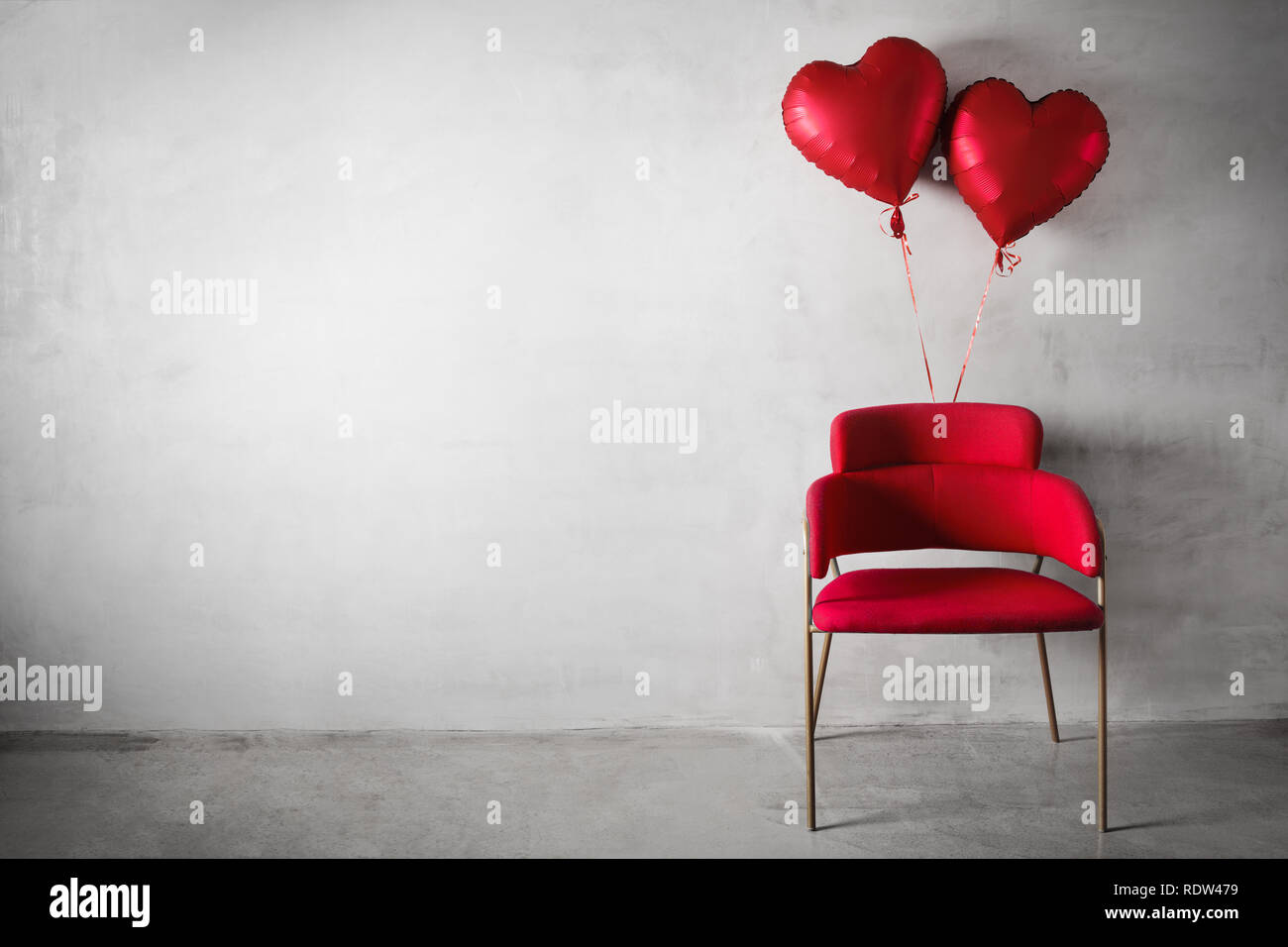 Silla Roja en forma de corazón con globos flotando sobre el muro de hormigón de fondo. Foto de stock