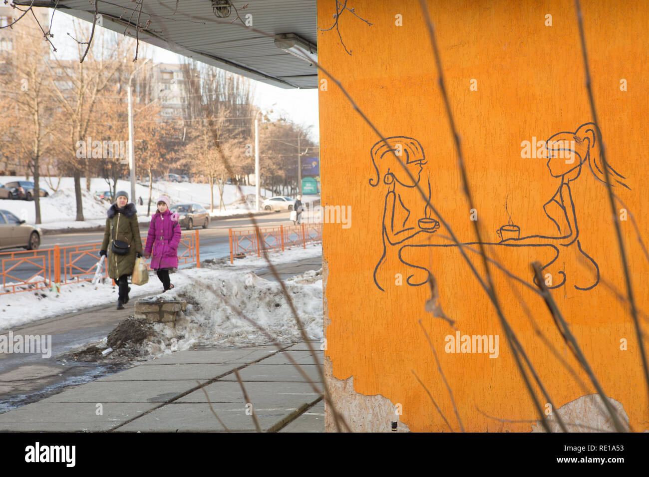 2 damas a pie de calle de Kiev un día de invierno, pasando por la pequeña tienda con elegante dibujo a mano graffiti de 2 damas bebiendo té caliente en una pared de la tienda amarilla Foto de stock