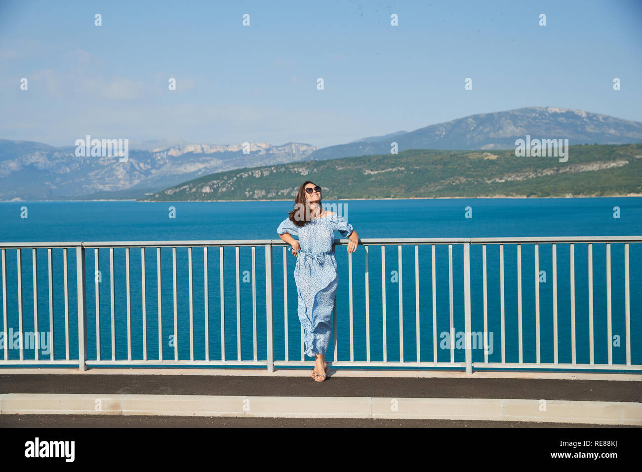 La hermosa chica en un vestido azul y gafas de sol posa sobre el puente, el largo cabello castaño, felices y sonrisas, el agua azul del lago y pendientes Foto de stock