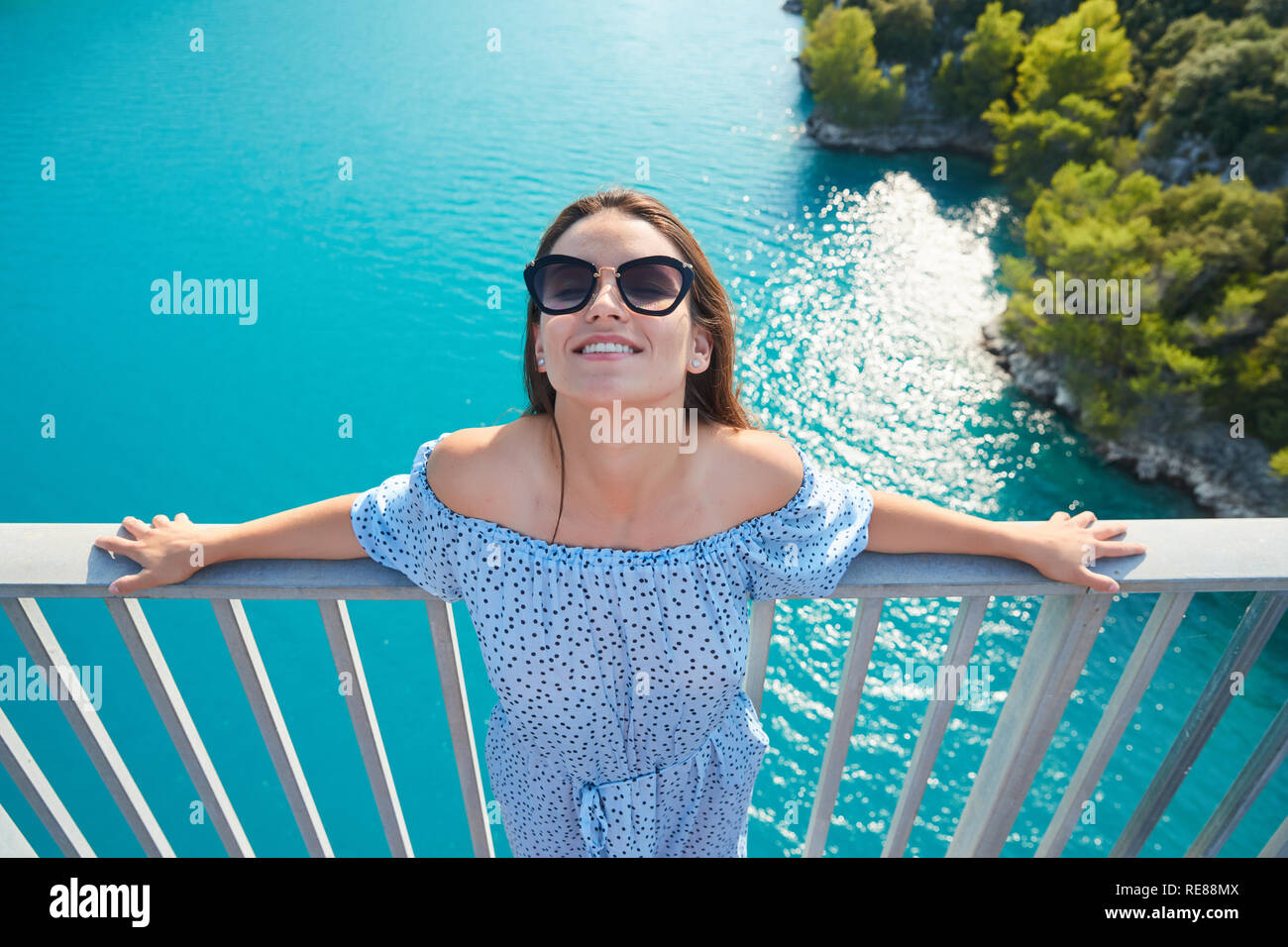 La hermosa chica en un vestido azul y gafas de sol posa sobre el puente, el largo cabello castaño, felices y sonrisas, el agua azul del lago y pendientes Foto de stock