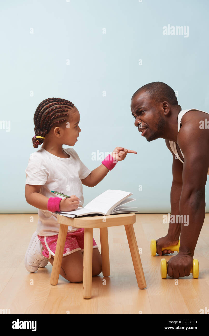 Cute little hija fitness su guapo negro padre joven, haciendo notas. Juego de rol de niño. Él la mira, escucha los reproches mientras ella Foto de stock