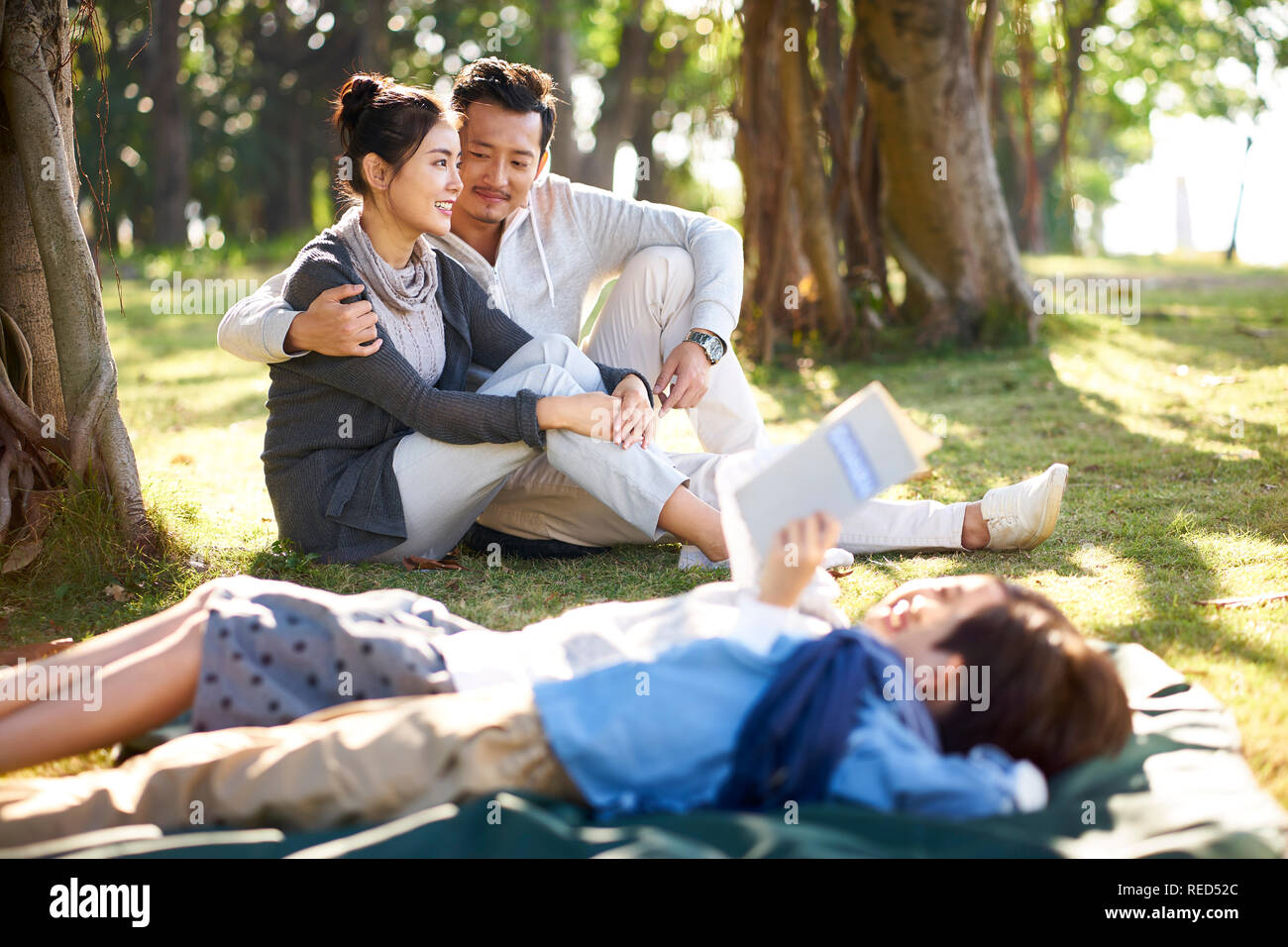 Pareja joven asiático sentados sobre el césped en el parque conversando con dos niños mentir libro de lectura en primer plano. Foto de stock