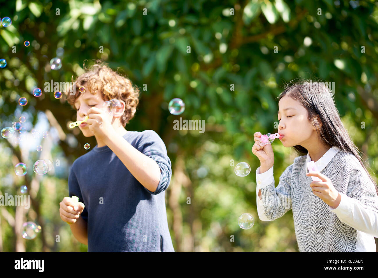 Little Girl asiáticos y caucásicos boy jugando juntos soplando burbujas de jabón al aire libre en un parque. Foto de stock