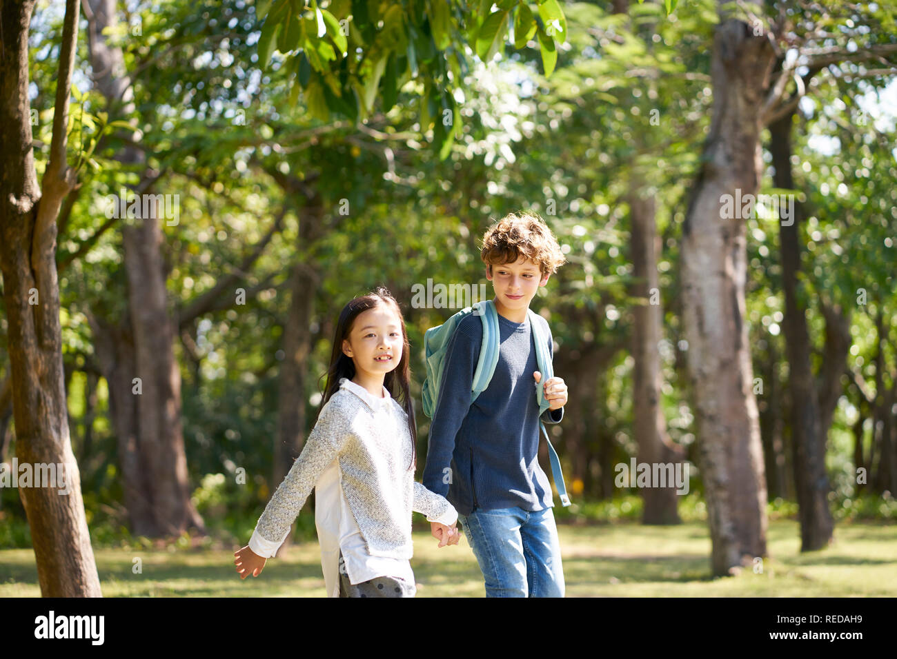 Little Girl asiáticos y caucásicos muchacho caminando juntos, tomados de las manos al aire libre en un parque. Foto de stock